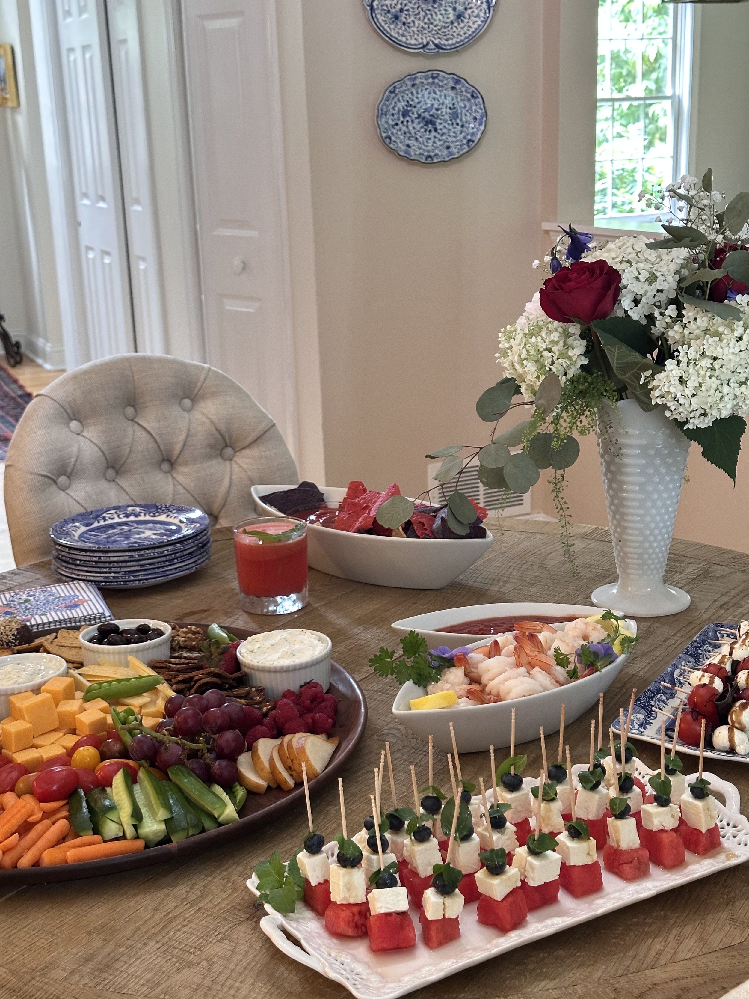 French-inspired Fourth of July entertaining table with a summer grazing board, watermelon feta skewers, shrimp cocktail, caprese skewers, and a hydrangea arrangement in a milk glass vase.