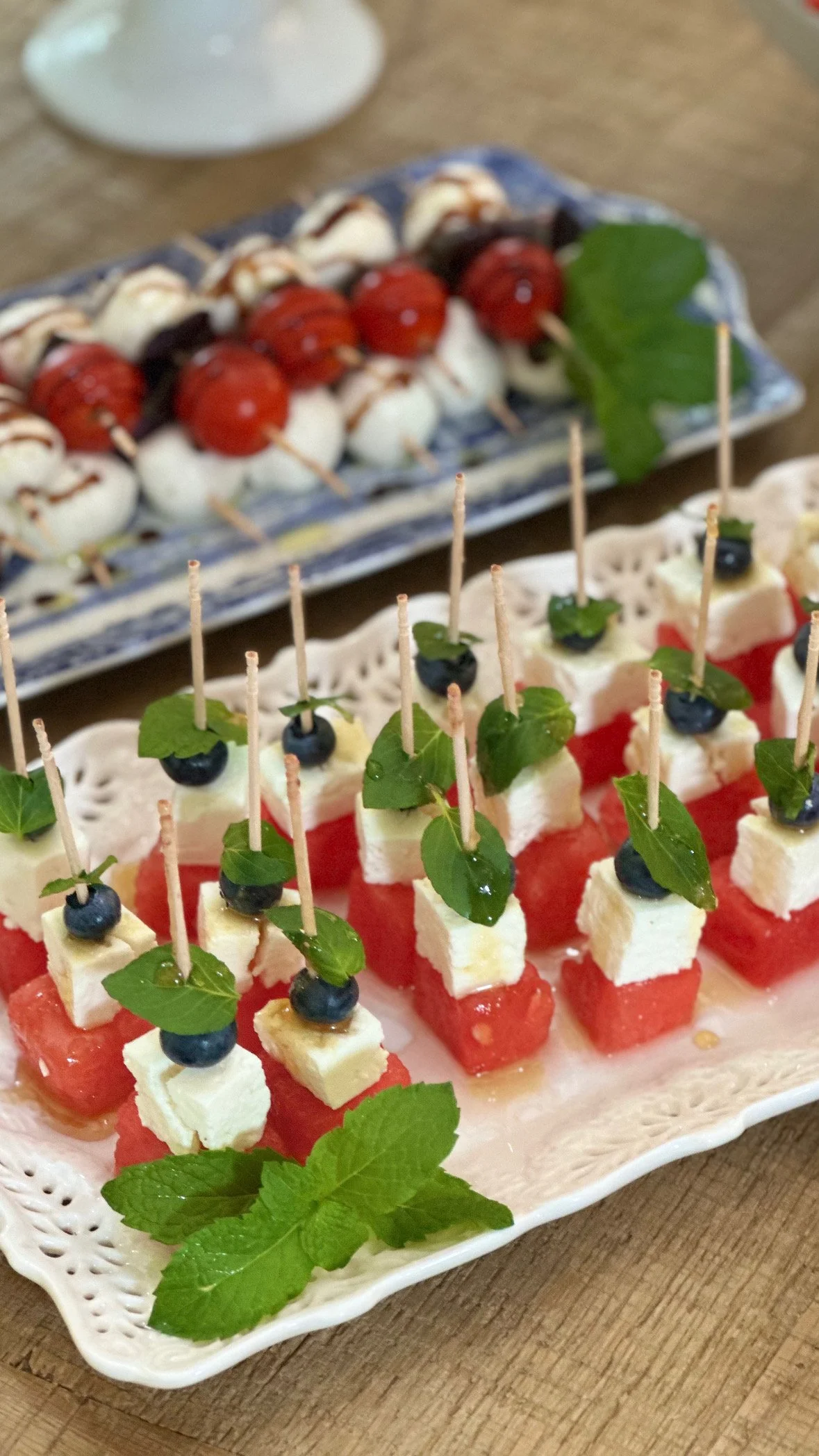 French-inspired Fourth of July appetizer spread with watermelon feta skewers on a white lace platter, caprese skewers on a blue and white platter, and a summer charcuterie board with cheese, fruit, and crackers.