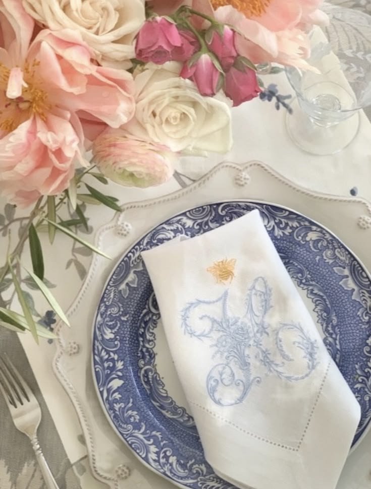 An embroidered white linen napkin with a blue monogram and gold bee, resting on blue and white transferware with a scalloped charger and peonies.