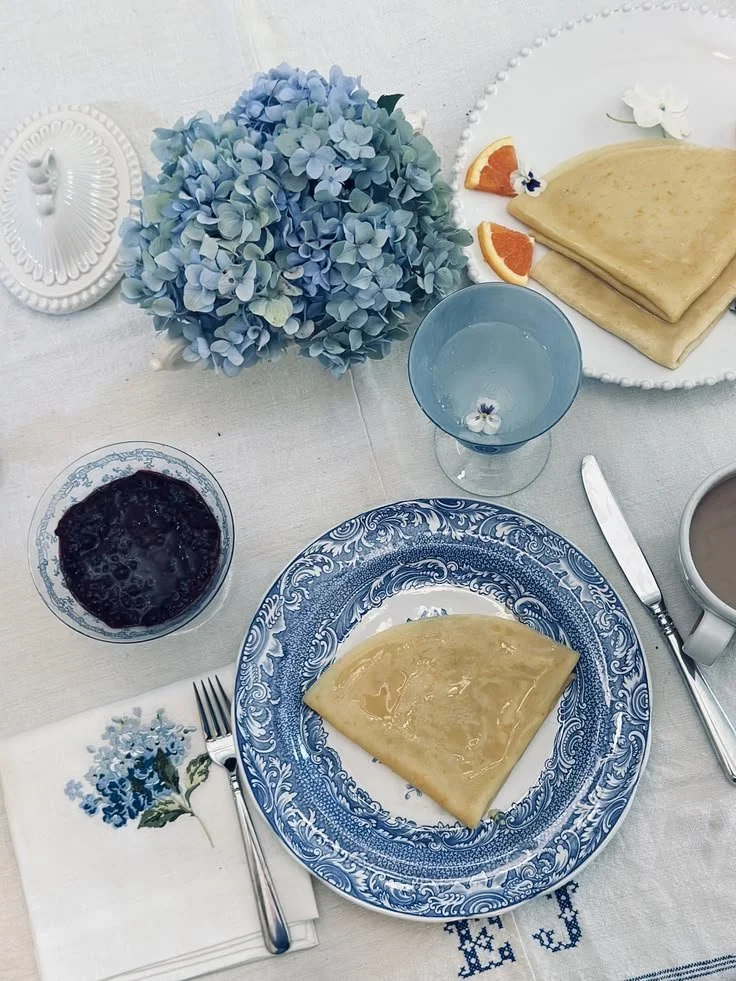Overhead flat lay of a French crêpe recipe with berry compote, blue and white transferware, and hydrangeas in a cream tureen