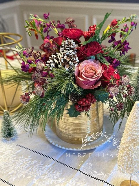 Seasonal centerpiece with red and pink roses, winter berries, evergreens, and a gold vase styled for a Christmas table.