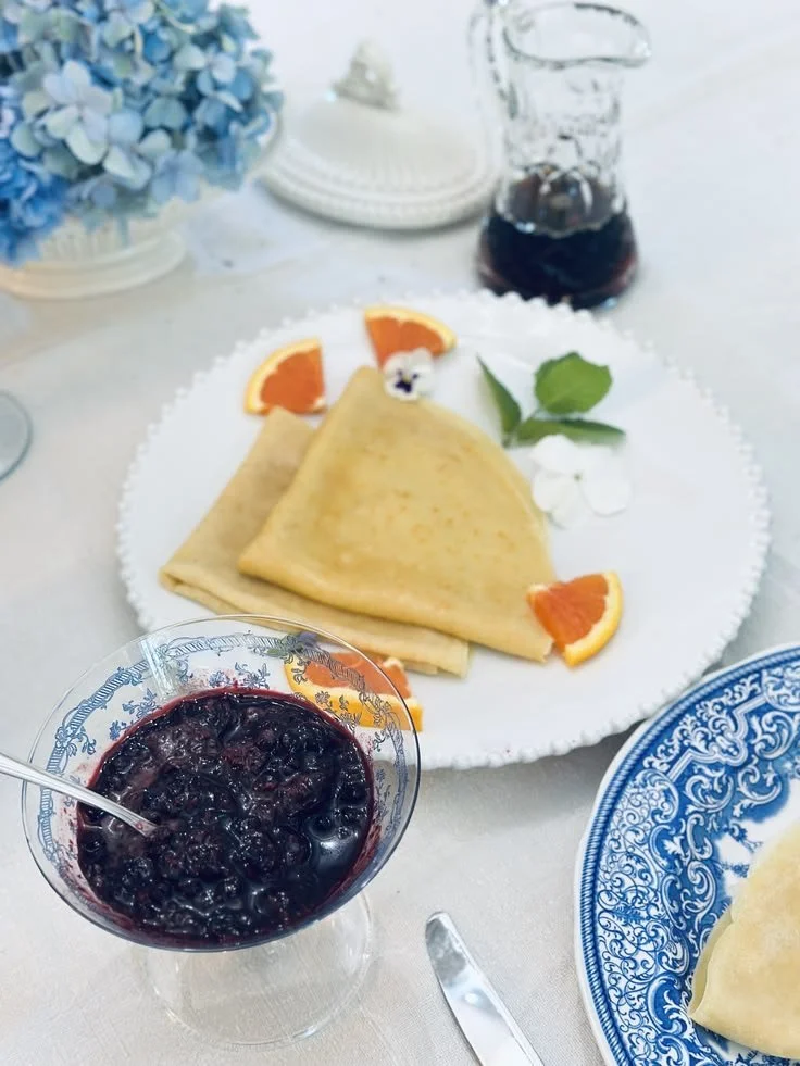 Folded crêpes with orange segments beside a crystal pitcher of warm Connecticut maple syrup