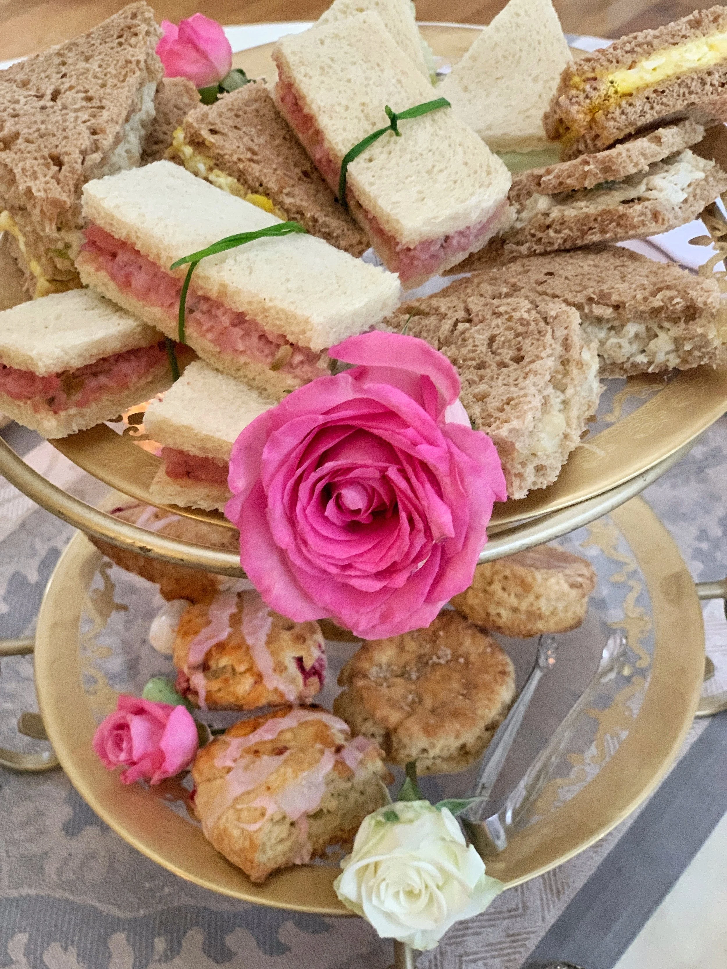 Elegant two-tiered serving stand with gold etched glass plates displaying tea sandwiches and raspberry scones for an Easter high tea gathering.