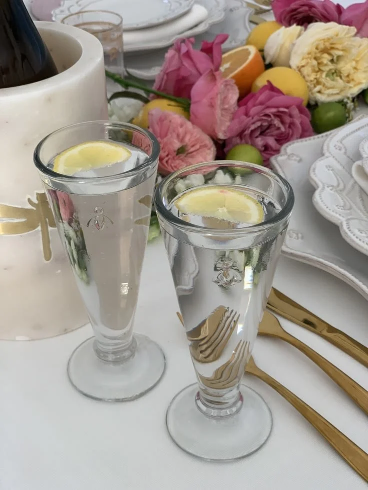 Set of French bee champagne glasses with a gold bee motif, filled with sparkling wine and lemon, on a summer table with roses and citrus.