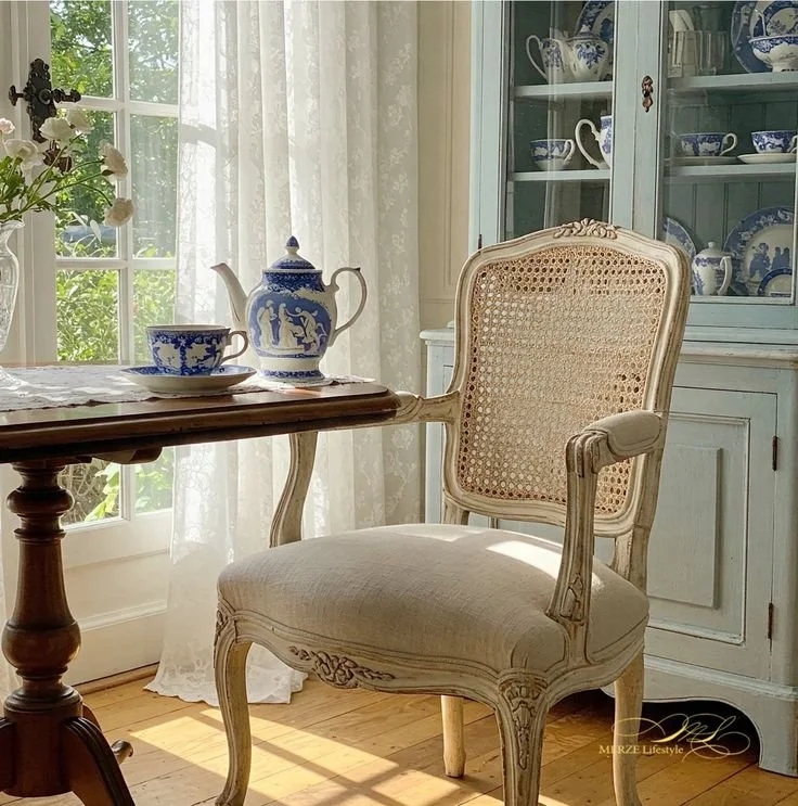 A kitchen with French chairs set near a table holding a blue and white tea set, with a hutch behind displaying matching porcelain, creating a space rooted in daily ritual and lived tradition.