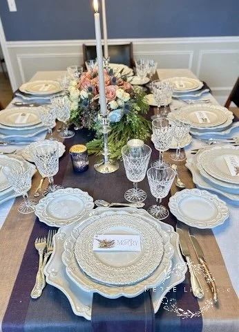 Angled view of a modern classic tablescape featuring cream layers, sterling silver flatware, Waterford crystal, woven linen runners, antique soup tureen centerpiece, and soft floral color.