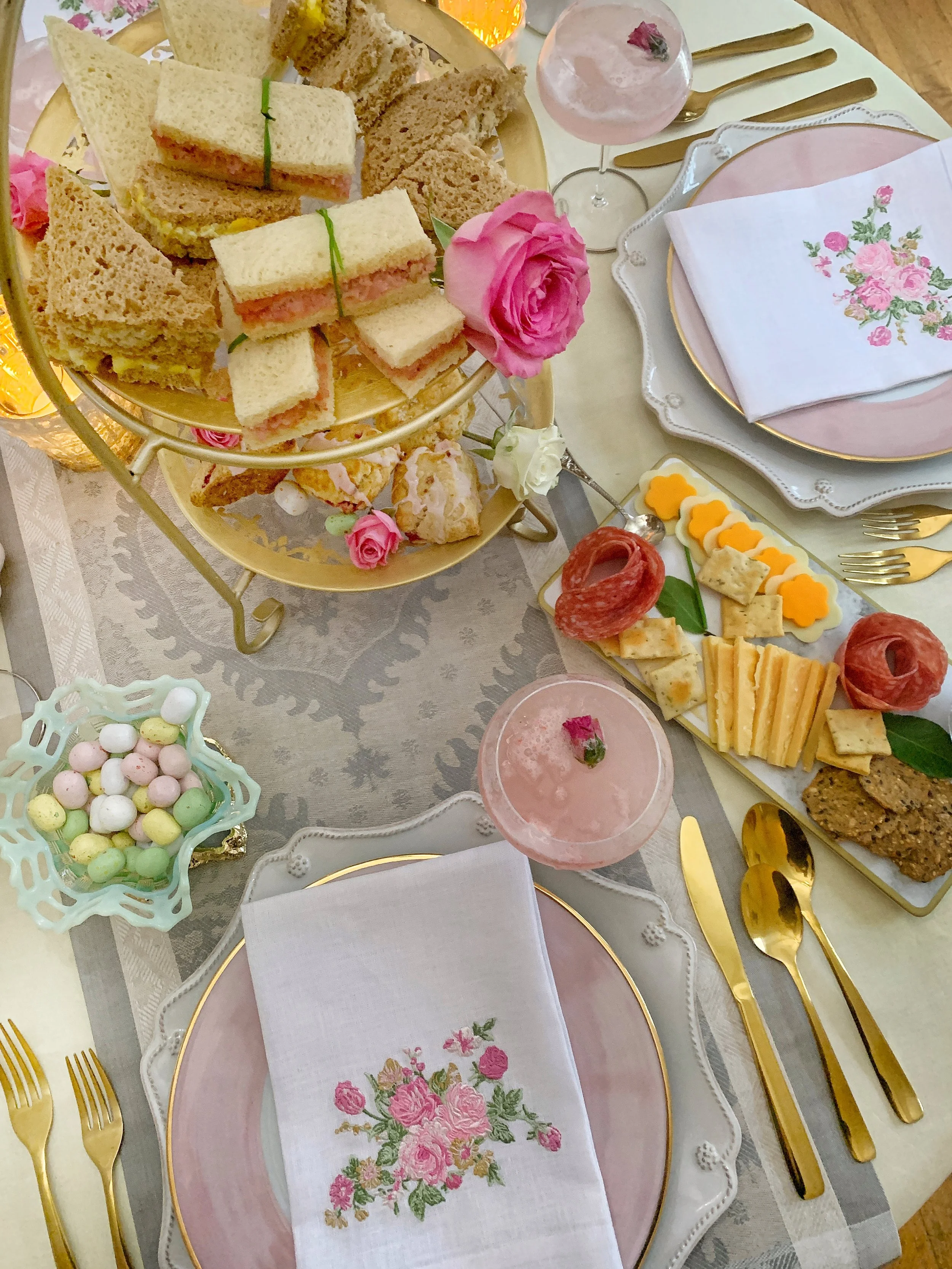Aerial view of an Easter high tea table featuring a charcuterie board, tiered tea caddy with finger sandwiches and scones, candy eggs, linen table runner, and etched glassware with a spring beverage.