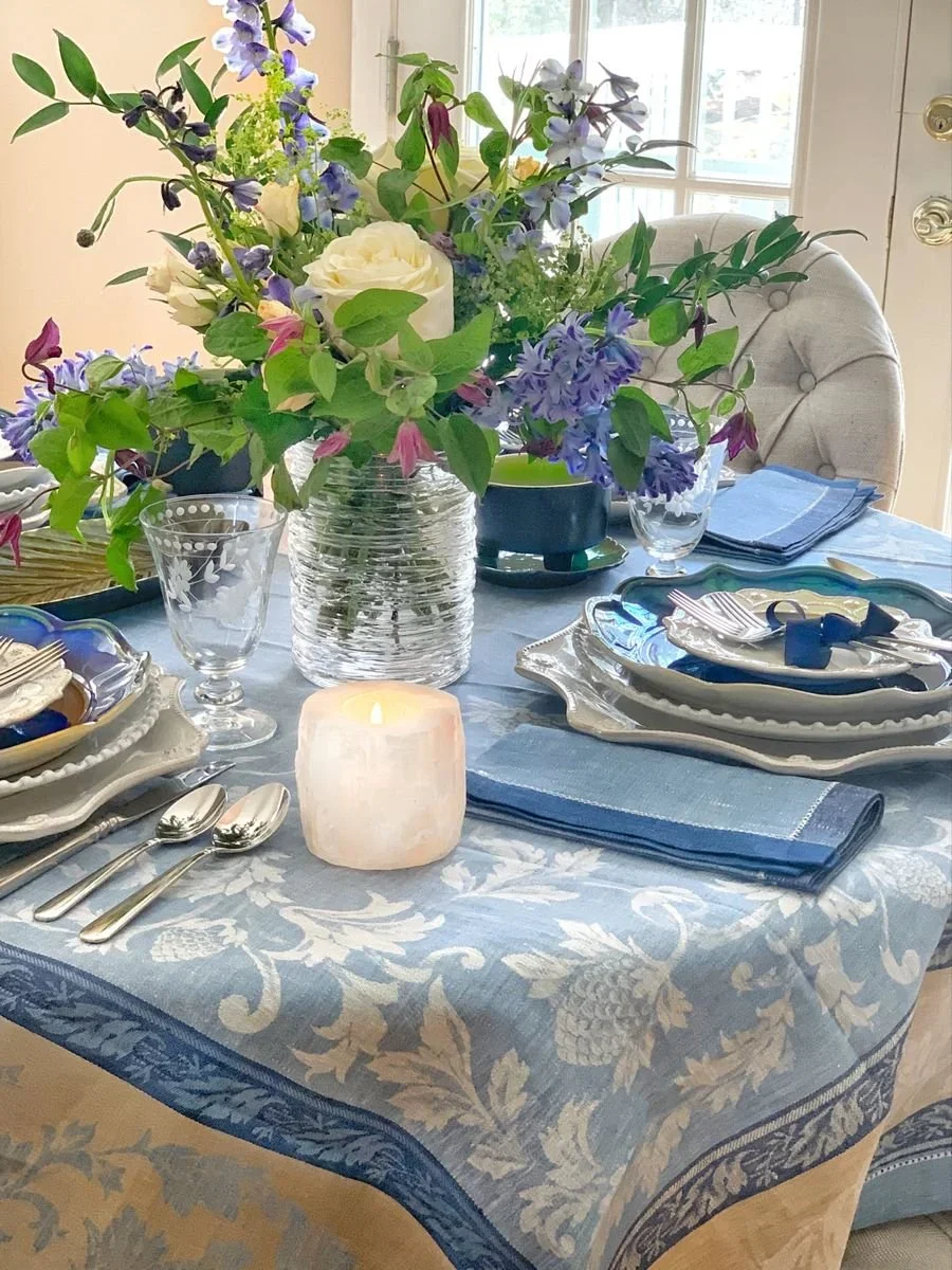 Full French coastal tablescape with a blue linen jacquard tablecloth, jewel-tone plates, candlelight, and a loose garden centerpiece
