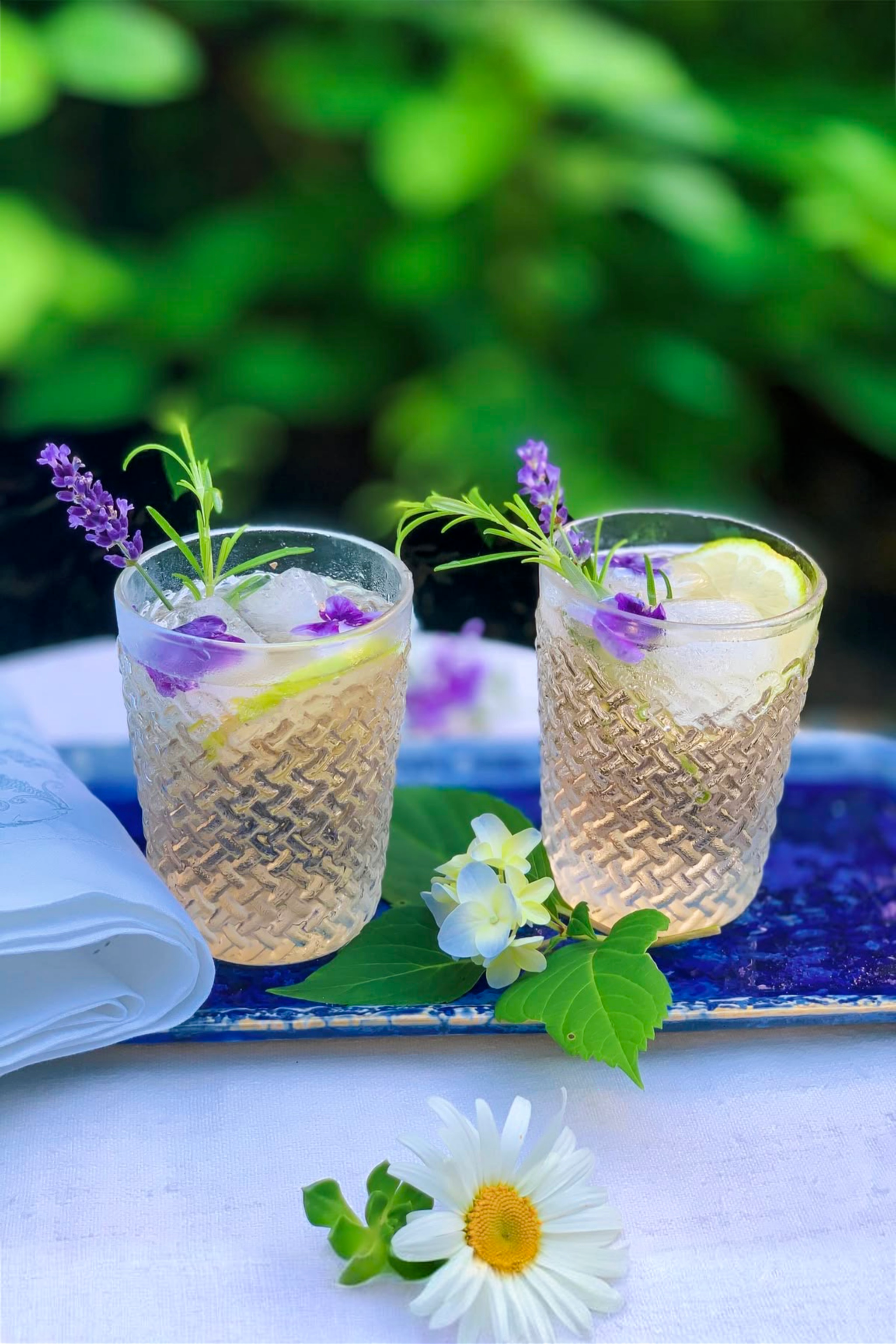 Garden drink garnished with edible flowers on a blue tray with daisies in the forefront.