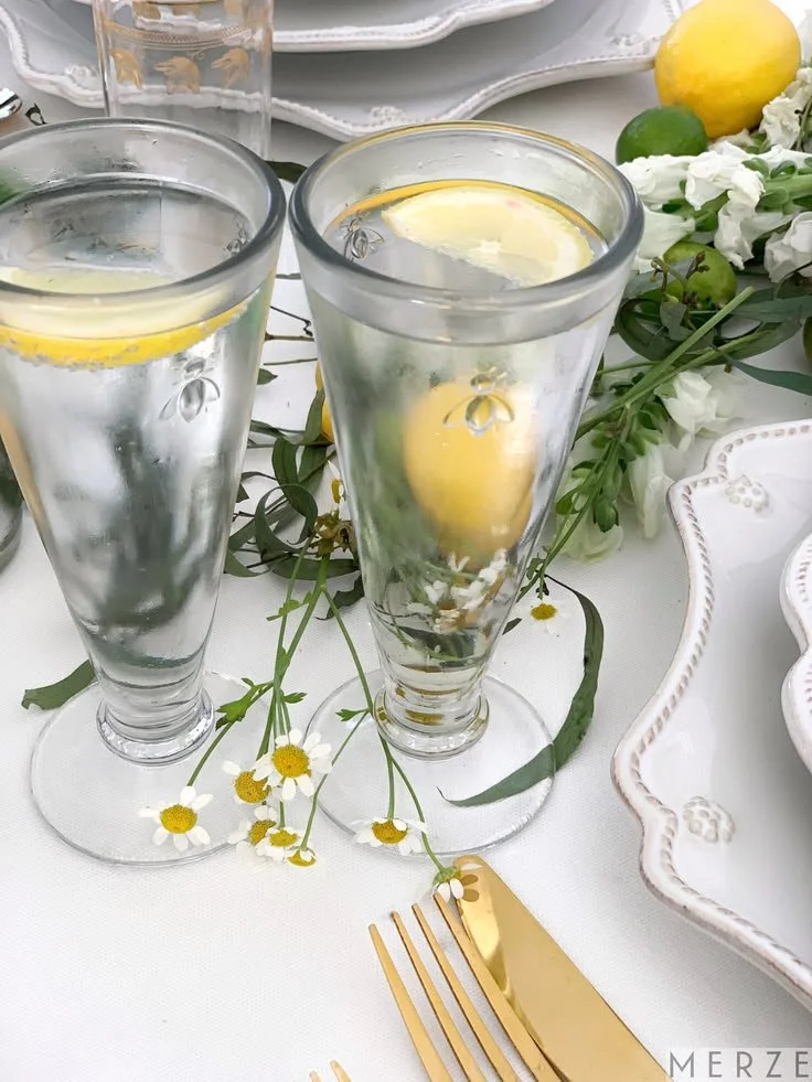 French bee champagne glasses with an etched bee detail and lemon, styled with chamomile and gold flatware on a white table.