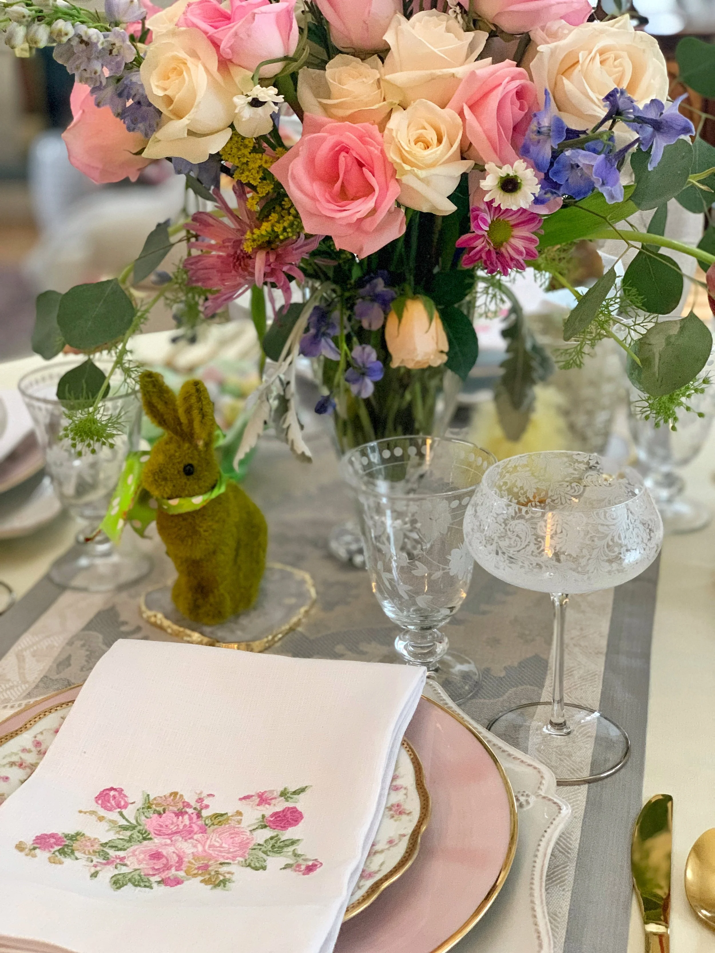 Spring Easter table place setting with moss bunny decoration, layered vintage china, etched glassware, and soft floral centerpiece.
