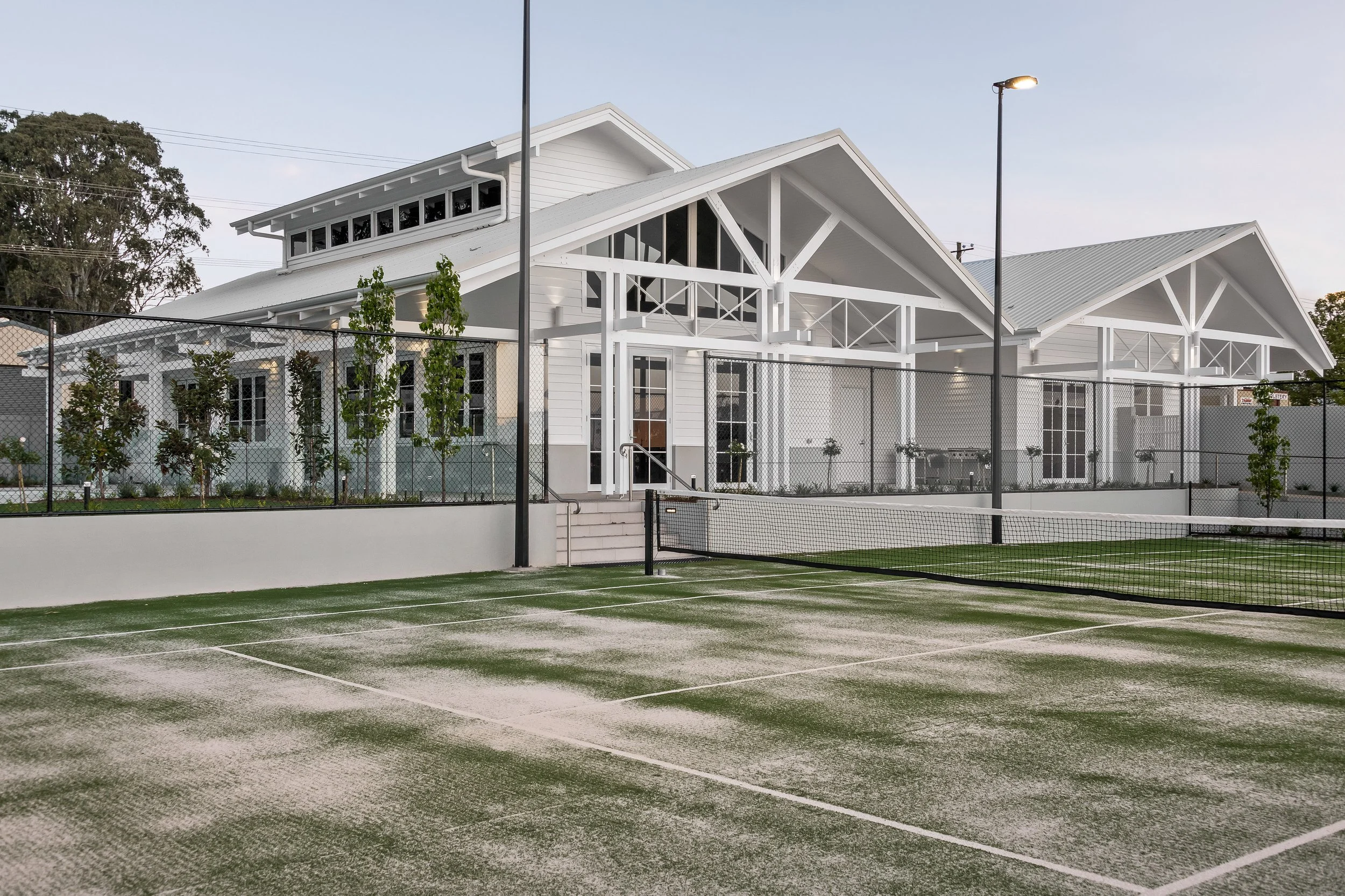 A tennis court with a green surface and white lines, surrounded by a chain-link fence, with a large white house with a covered porch in the background.