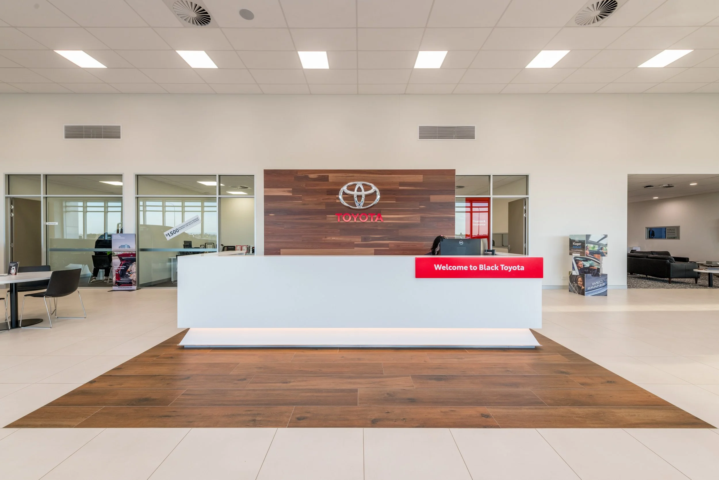 Black Toyo showroom reception area with a white counter and a wooden backdrop displaying the Toyota logo and the text 'Welcome to Black Toyota'. There are glass-walled offices behind the counter, and a waiting area with black sofas on the right side.