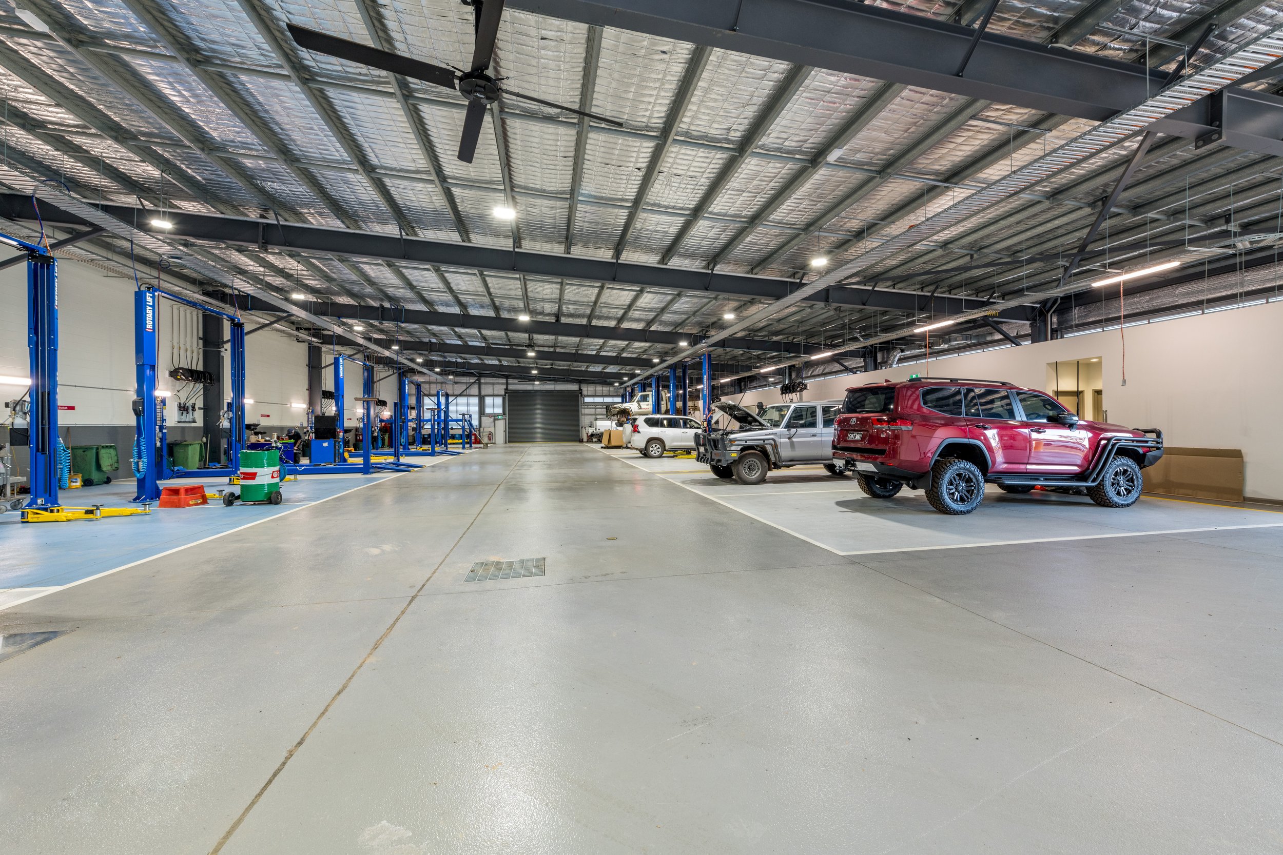 Black Toyota warehouse with a row of vehicle lifts on the left and several cars on the right, including a red off-road vehicle, a gray pickup truck, and a white car, with a high ceiling and industrial lighting.