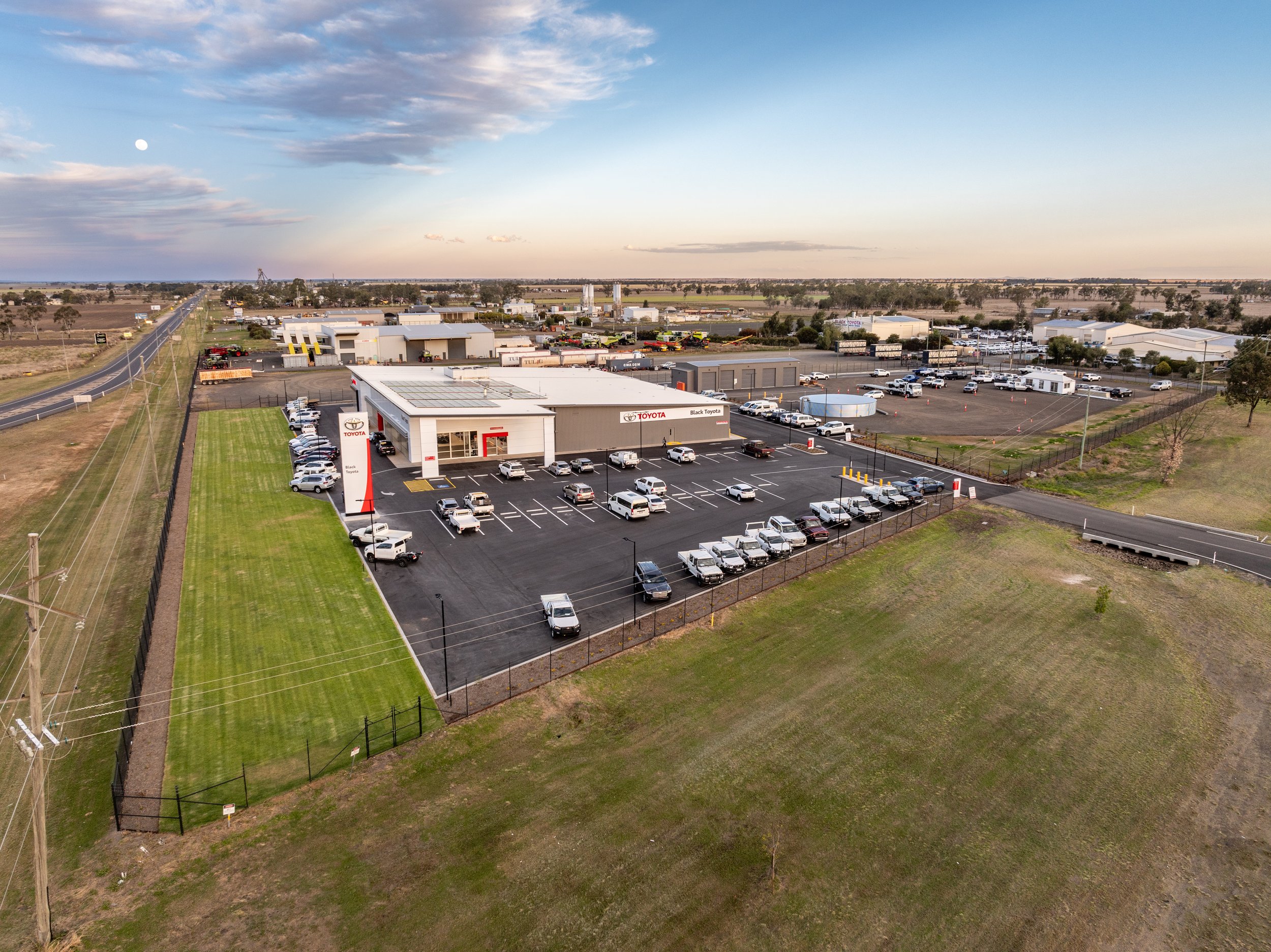 Aerial view of the Dalby Black Toyota car dealership with a large parking lot, surrounding grassy areas, and multiple buildings in the background, under a partly cloudy sky at dusk.