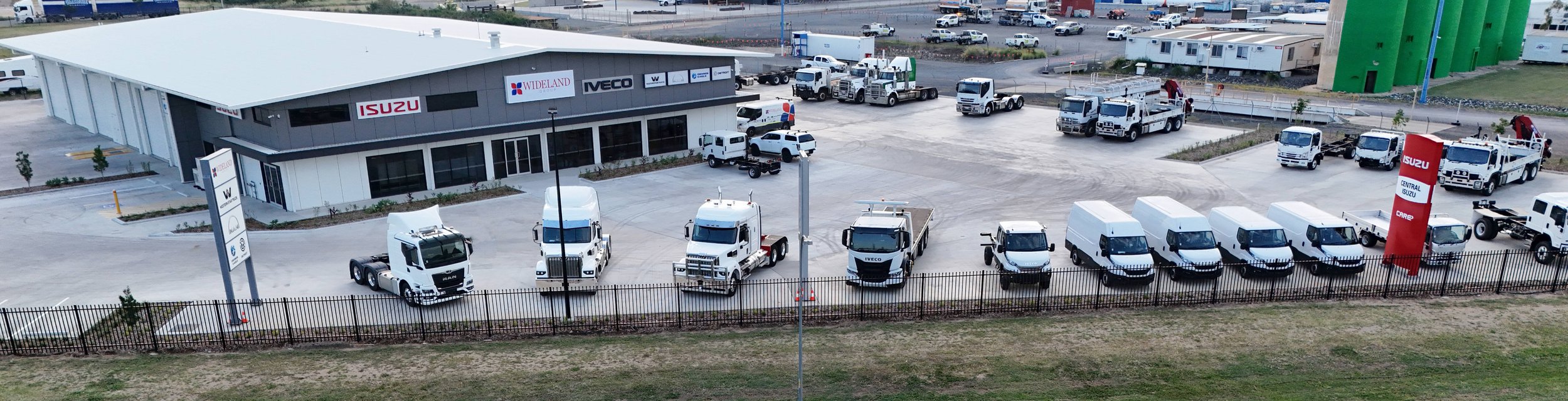 An aerial view of a car dealership lot with multiple white trucks and vans parked in front of a building with signs for Isuzu, Wideland, VECO, and other brands. There is a fenced perimeter and a parking lot with other vehicles in the background.