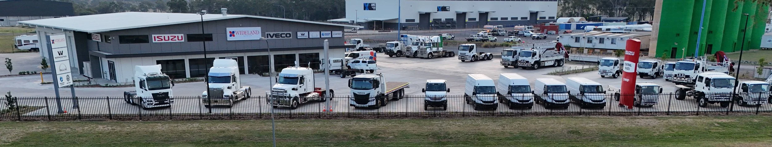 A vehicle dealership lot with white trucks and vans parked in front of a building that has signs for Isuzu and other brands.