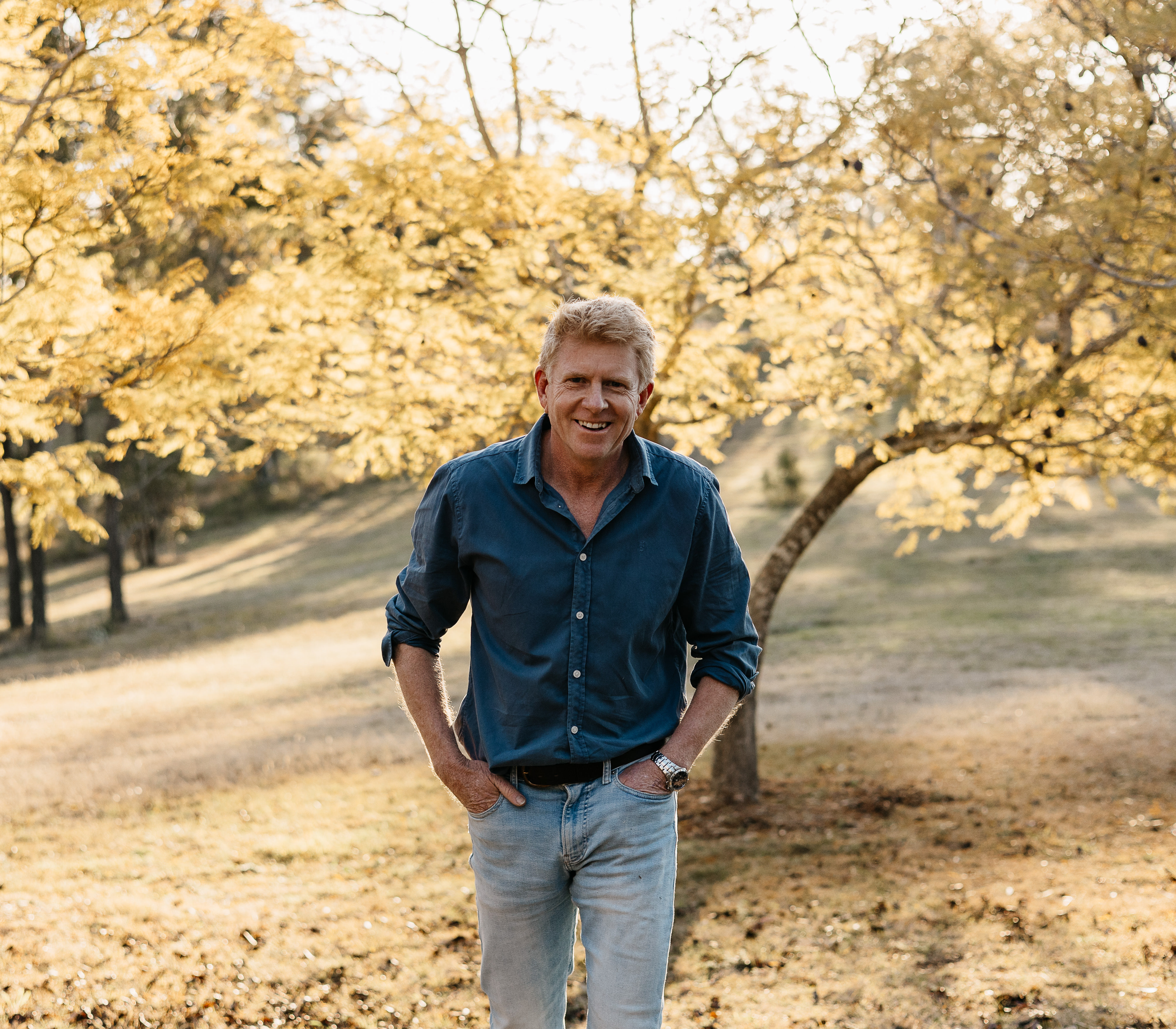 A smiling man in a blue shirt and jeans standing outdoors in front of a yellow-leaved tree in autumn.