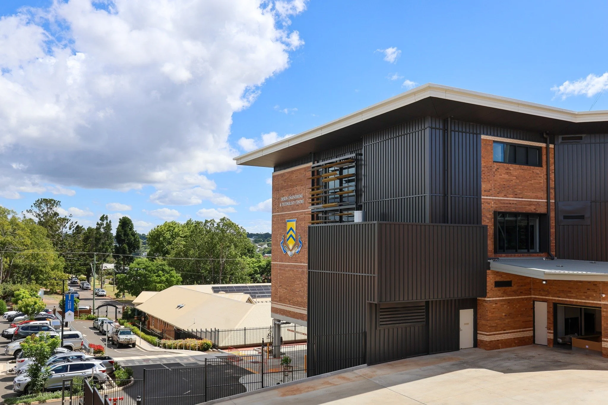 Modern brick and black metal building with sign for Design Engineering & Technology Center, parking lot with cars, trees, and partly cloudy sky.