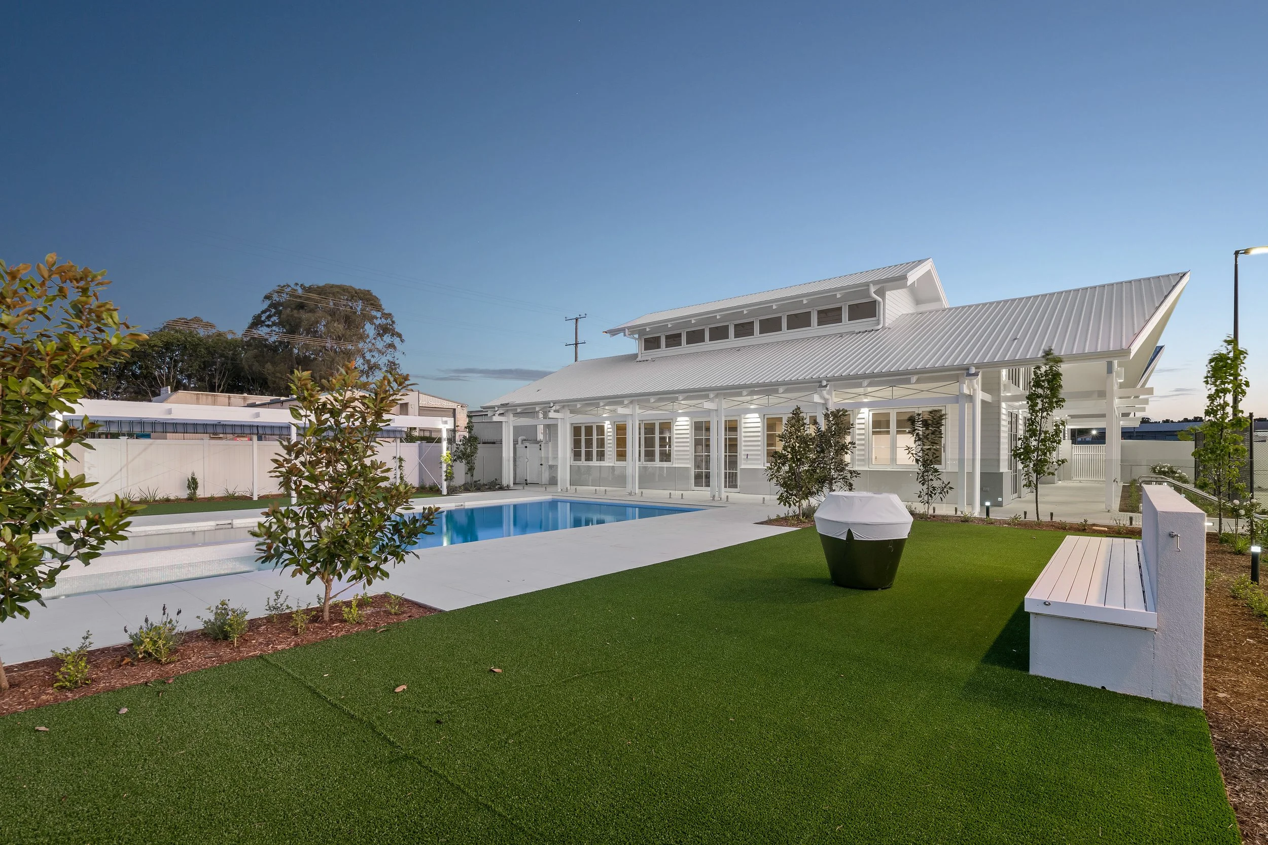 A modern house with a white exterior and a gabled metal roof, a swimming pool, and a well-manicured lawn with small trees and plants, taken at dusk.