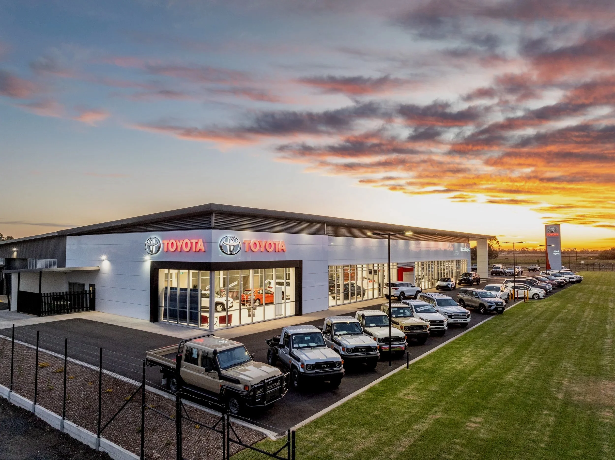 An exterior view of a Toyota dealership at sunset, showing a modern building with large glass windows, Toyota logos illuminated on the facade, a row of parked cars including trucks and SUVs, and a grassy area with a few light posts.