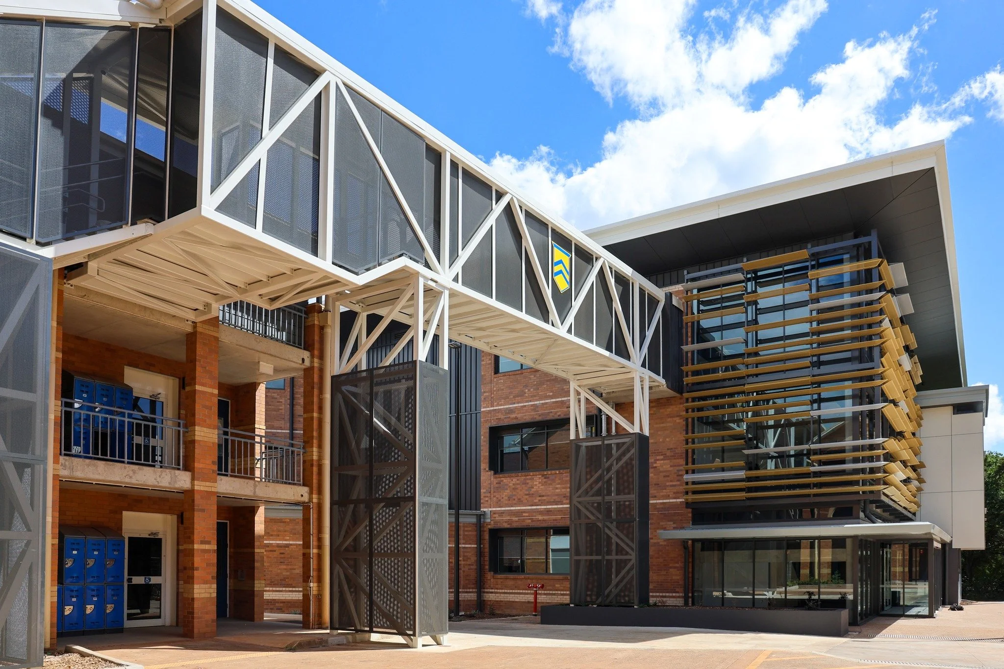 Modern multi-story building with brick walls, glass windows, metal and wooden exterior design, including a covered entrance and an external staircase, under a blue sky with white clouds.