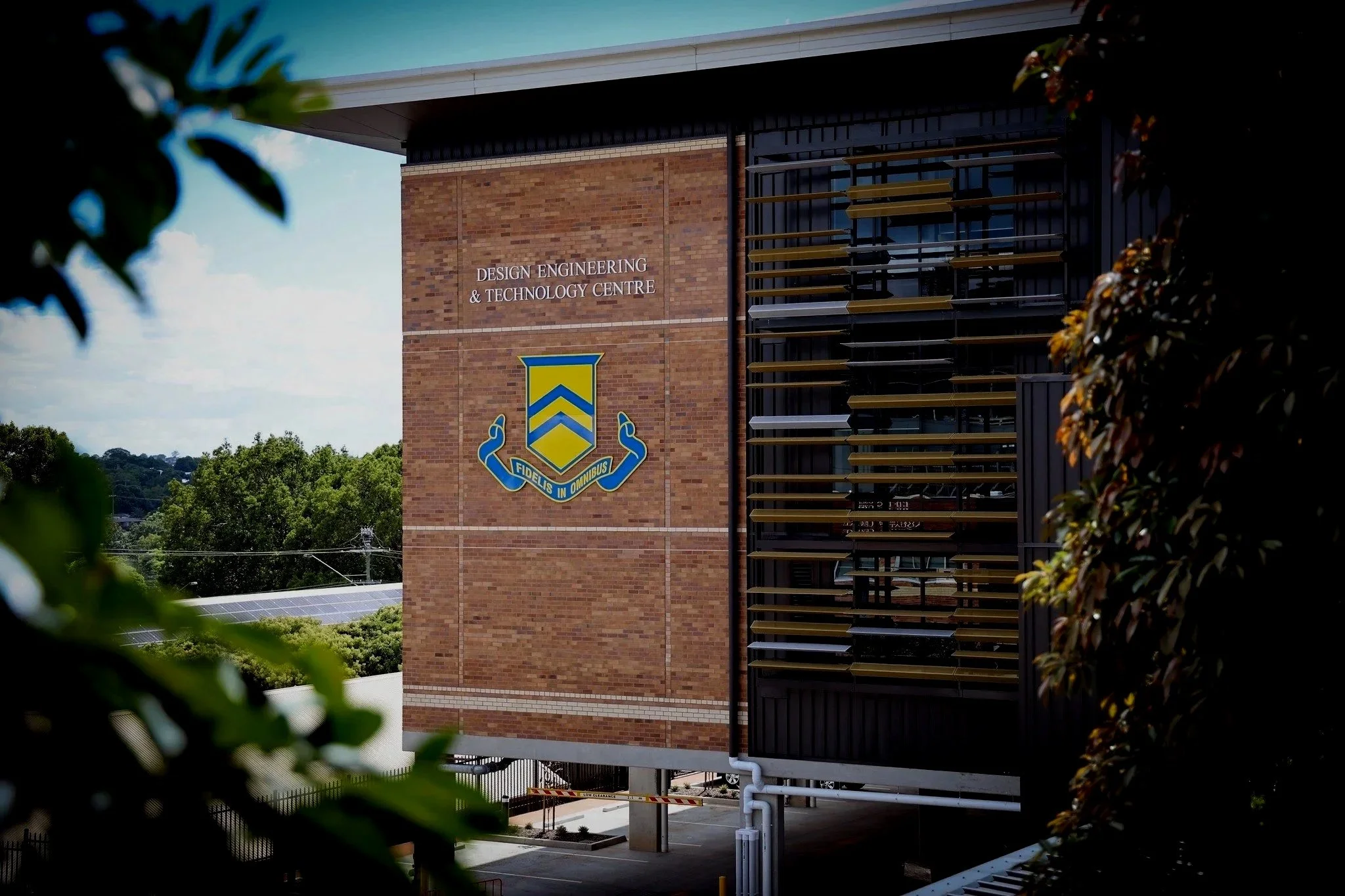 Exterior view of the Design Engineering & Technology Centre building with a brick wall, TGS school emblem, and a window with horizontal black and gold shading panels.