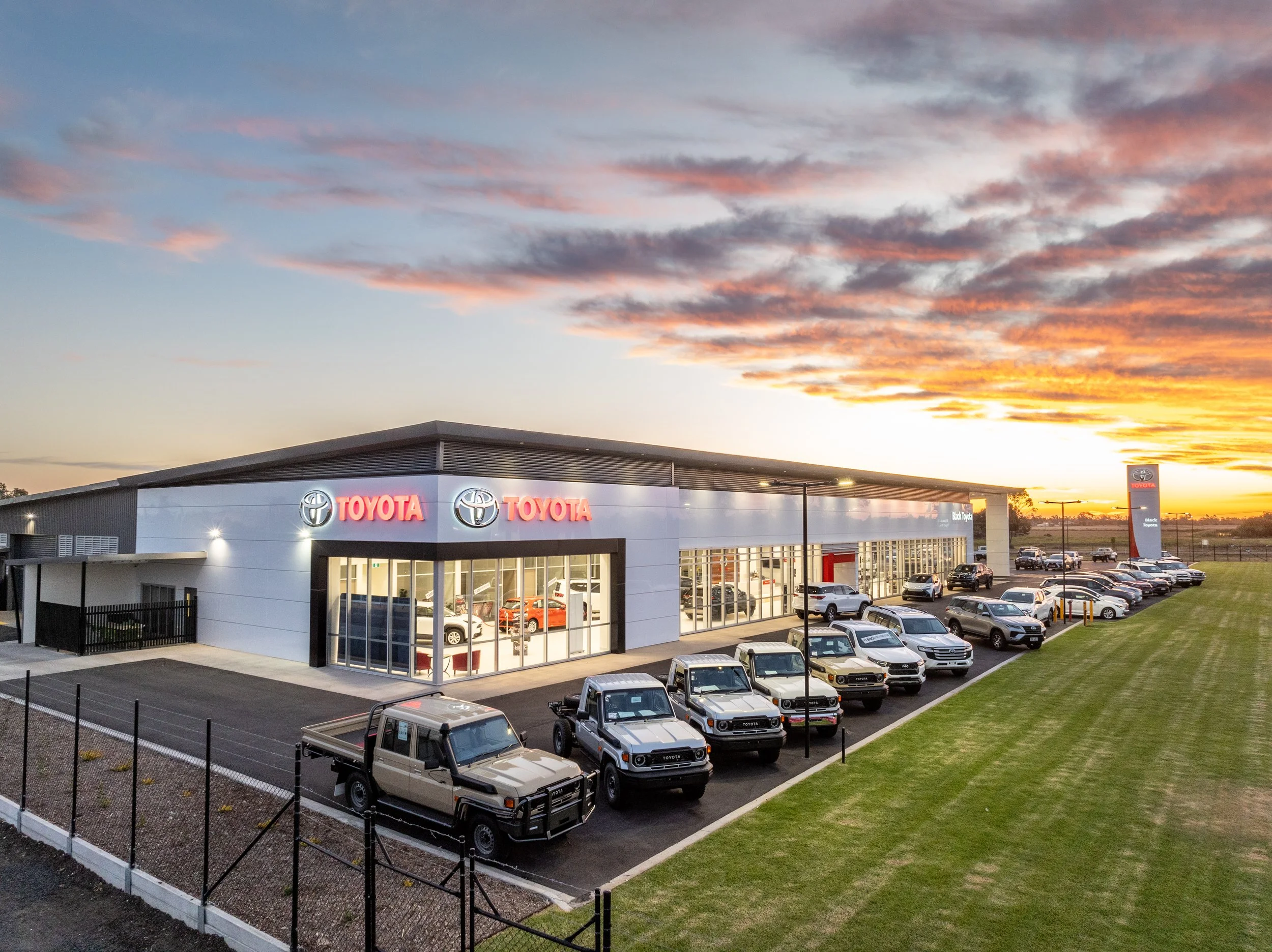Exterior view of the Black Toyota car dealership at sunset, with a row of parked vehicles in front and a large sign in the background.