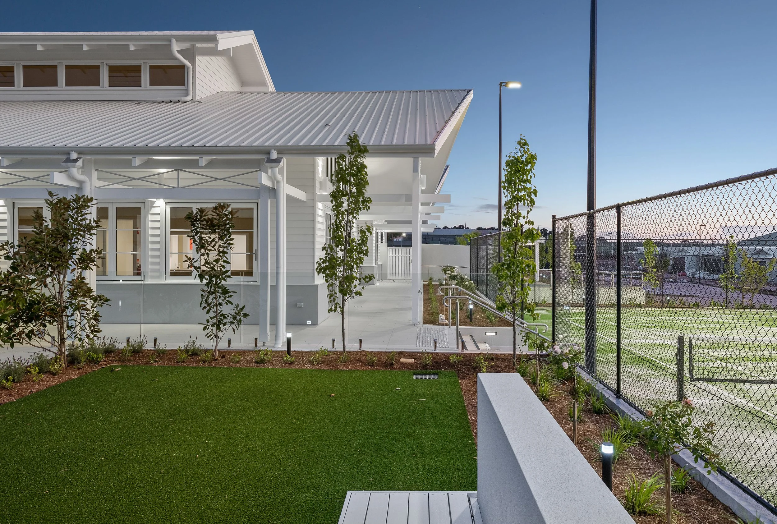 Modern building with white exterior walls, large windows, and a metal roof, surrounded by landscaped yard with grass, small trees, outdoor lighting, and a chain-link fence enclosing a sports court, during early evening or dusk.