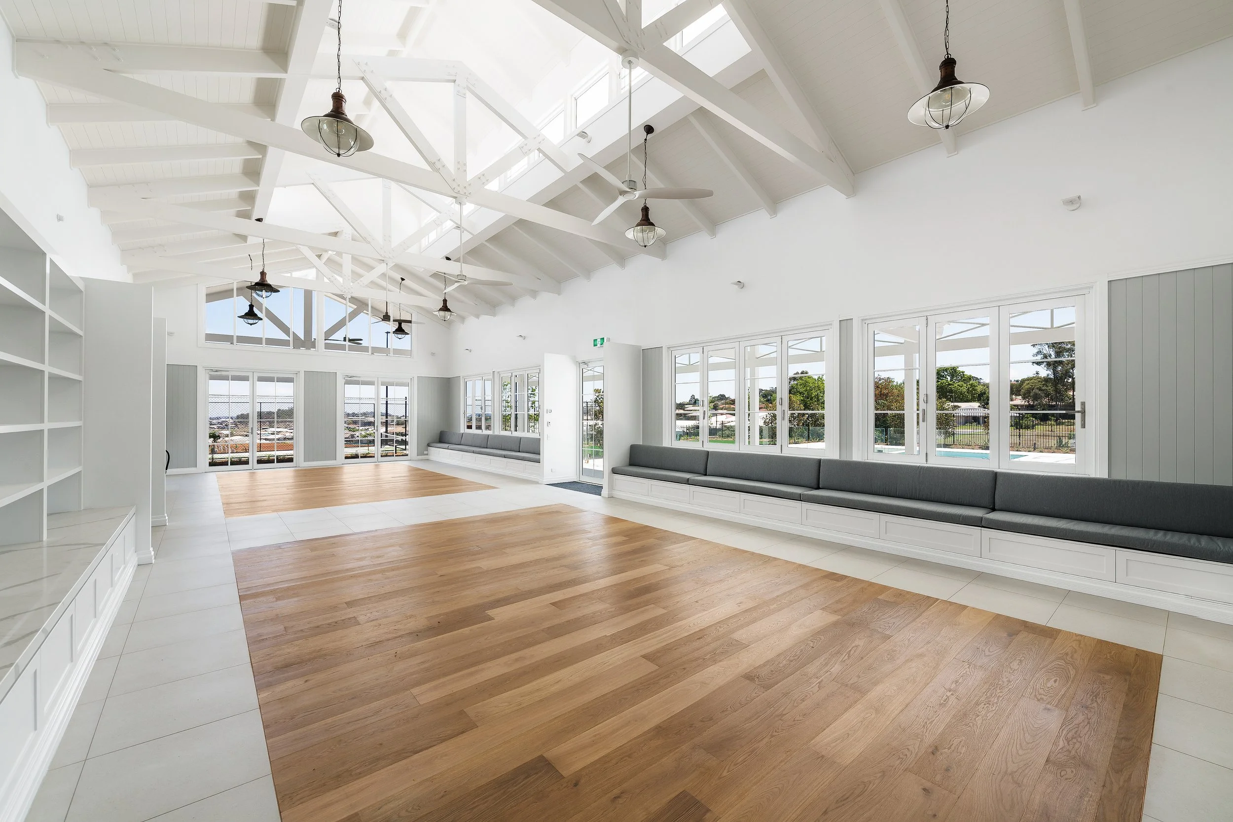 Spacious room with white walls, high beamed ceiling, large windows, gray cushioned built-in benches, and a mix of wood and tile flooring.