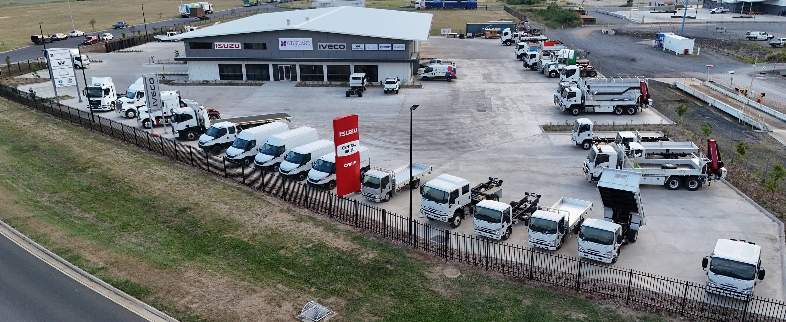 Aerial view of a commercial vehicle dealership lot with various trucks, vans, and service vehicles parked in front of a building displaying signs for Isuzu and Wideland.
