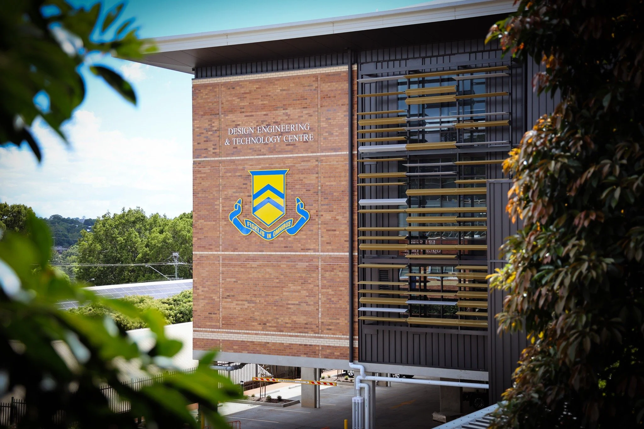 Exterior view of the Design Engineering & Technology Centre building with a brick facade, TGS school emblem, and modern window louvers, surrounded by trees and greenery.