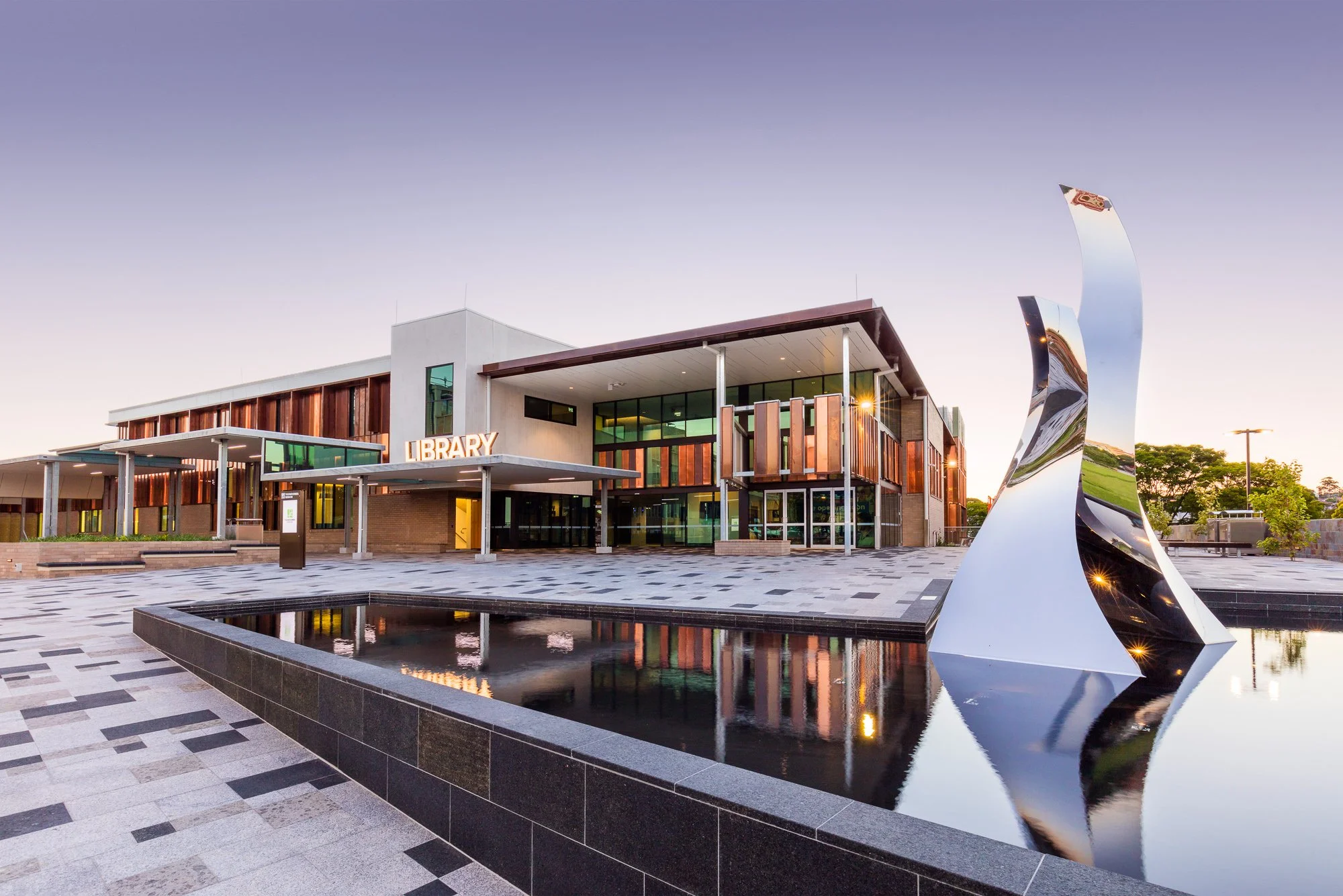 Modern library building with glass windows and wooden accents, reflecting in a water feature, during sunset.