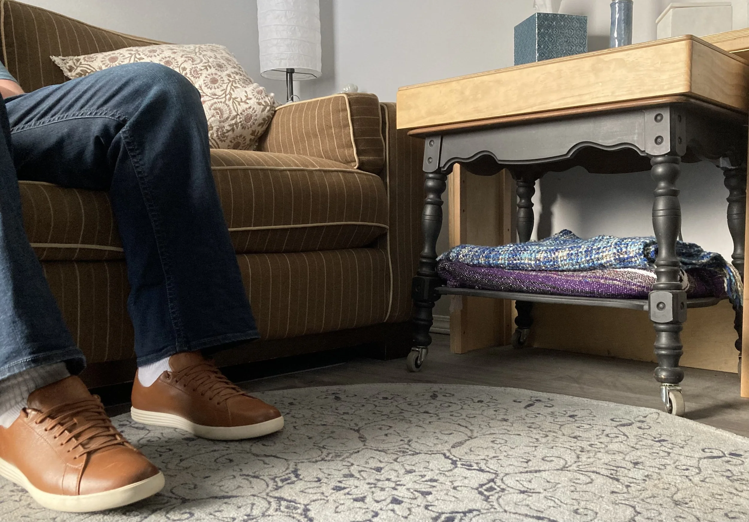 A room with a brown striped sofa, a patterned pillow, a wooden side table with a blanket underneath, a small lamp with a white shade, and part of a person wearing dark jeans, white socks, and tan sneakers sitting on the sofa.