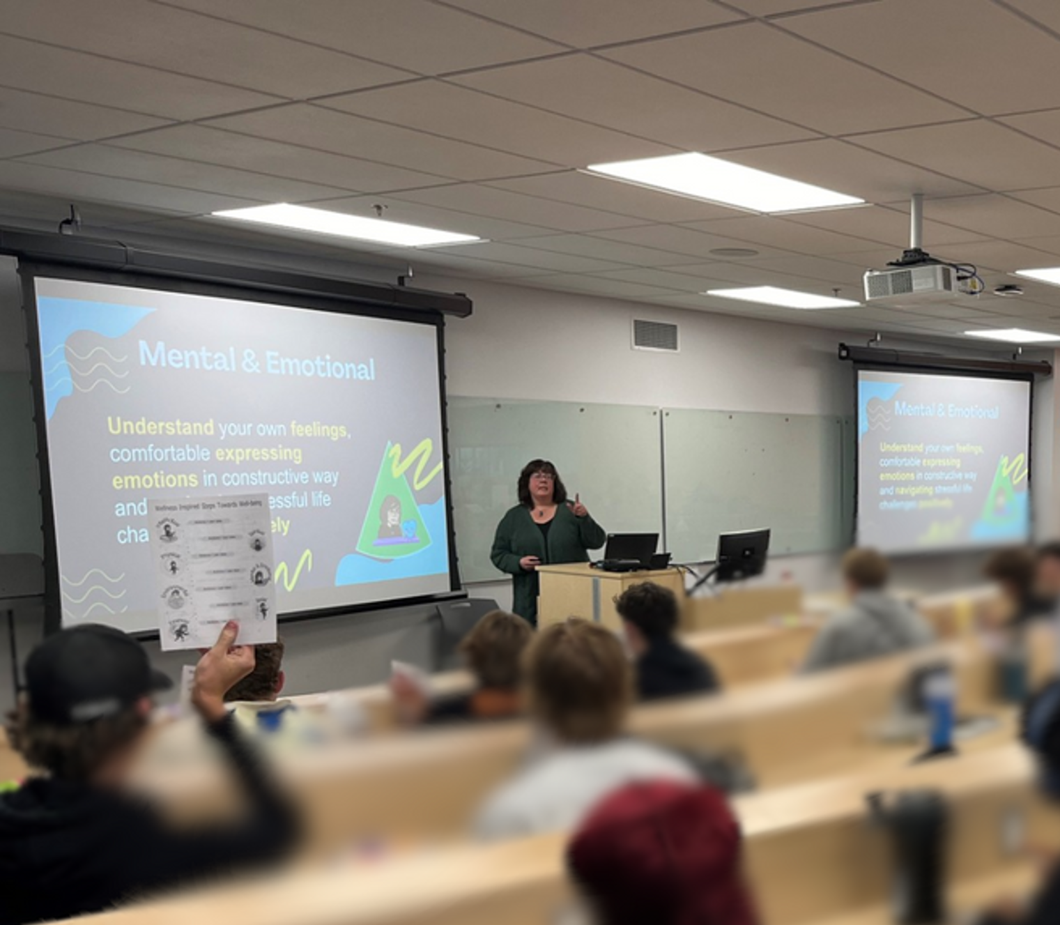 A classroom setting with a woman presenting on mental and emotional health. Multiple students are seated, some taking notes or raising hands. Two screens display educational slides on the topic.
