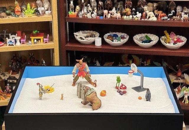 Display of figurines in a sand tray, including a large a bear, a basketball hoop, a penguin, a knight and various other animal and human figures, with shelves of additional figurines in the background.