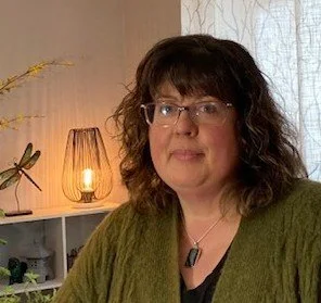 A woman with shoulder-length dark curly hair and glasses, sitting indoors in front of a light-colored wall with a lamp and a plant in the background.
