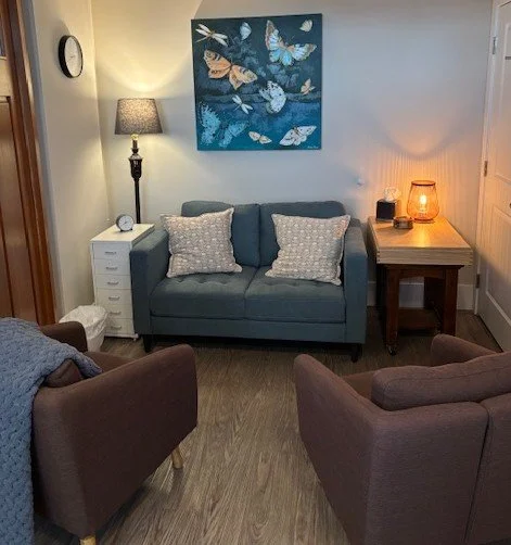 Counselling office with a blue loveseat, two armchairs, a table lamp, a small white cabinet, a side table with a lamp, and butterfly artwork on the wall.