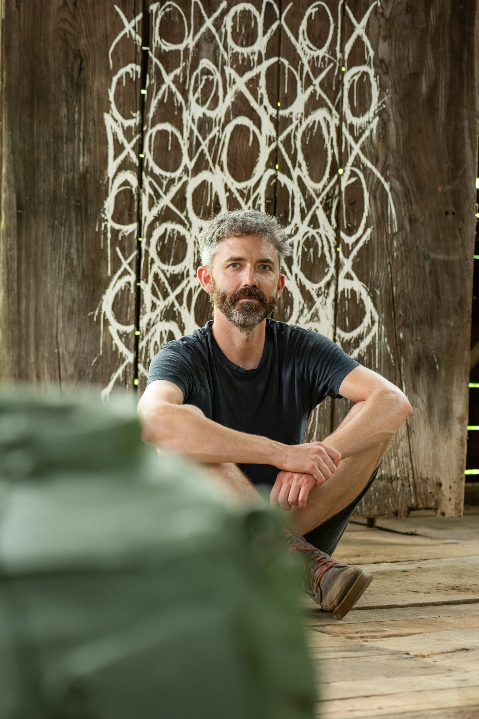 A sitting white bearded man with graying hair with a green pot blurred in front against a brown wall with circles