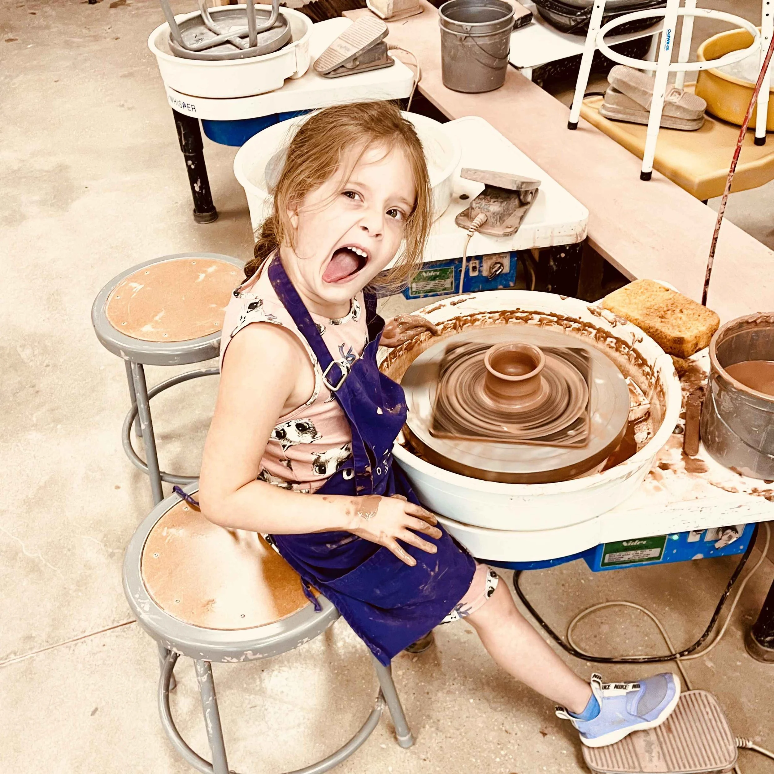 a young girl sticking her tongue out while throwing on the pottery wheel