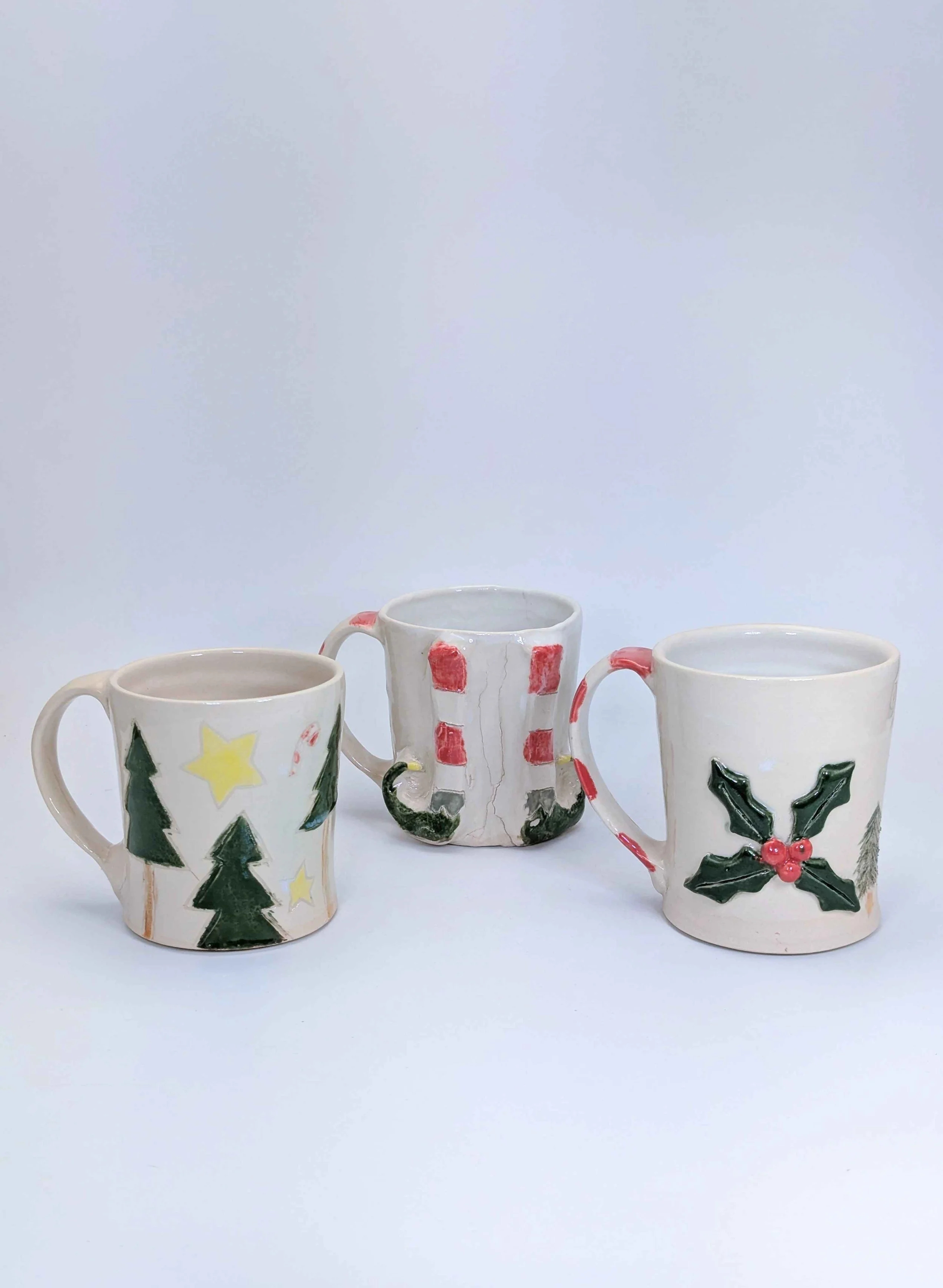 Three decorated christmas mugs with winter decor like trees, mistletoe and stockings