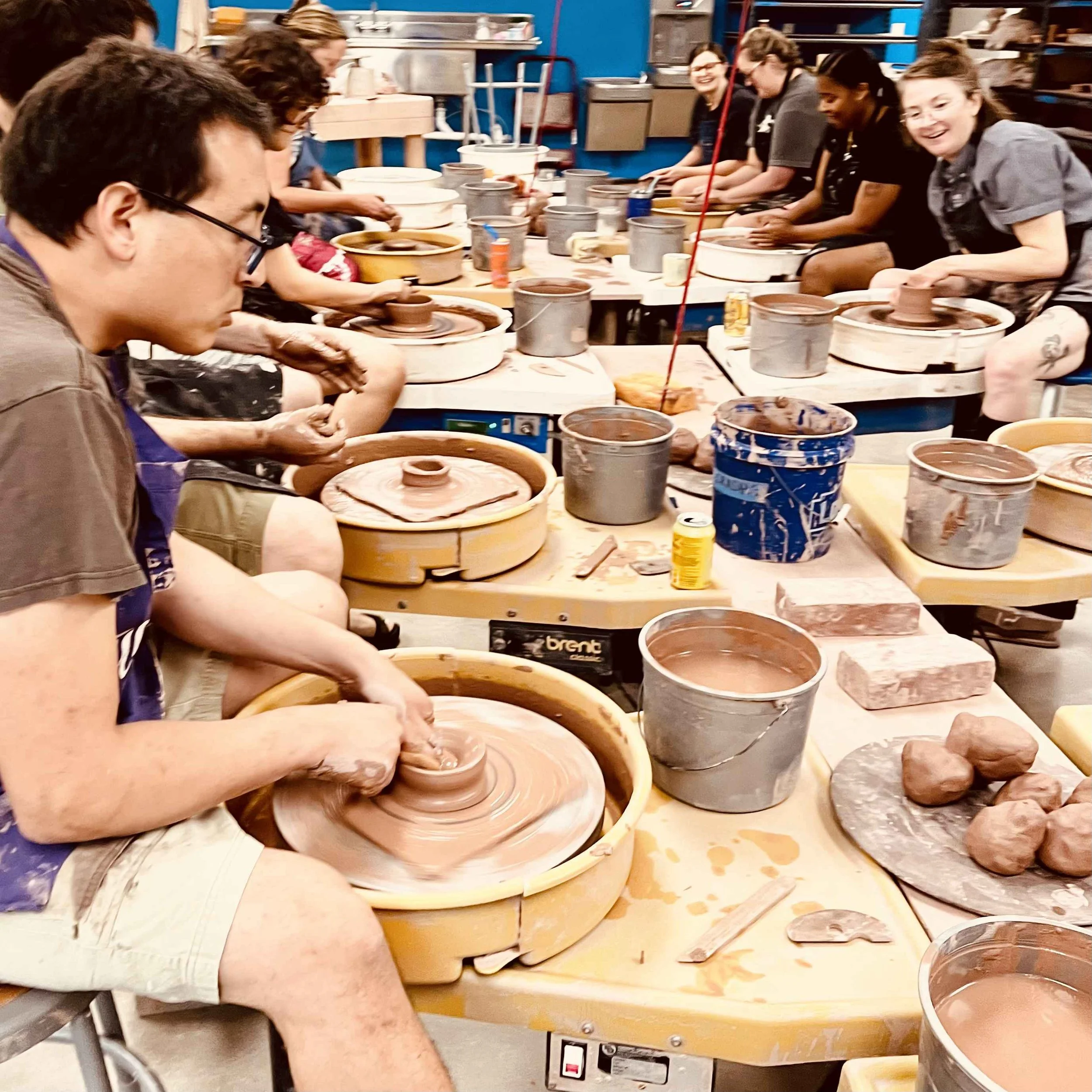Multiple Individuals sitting at pottery wheels in the Studio T/M classroom getting muddy and making on the wheel