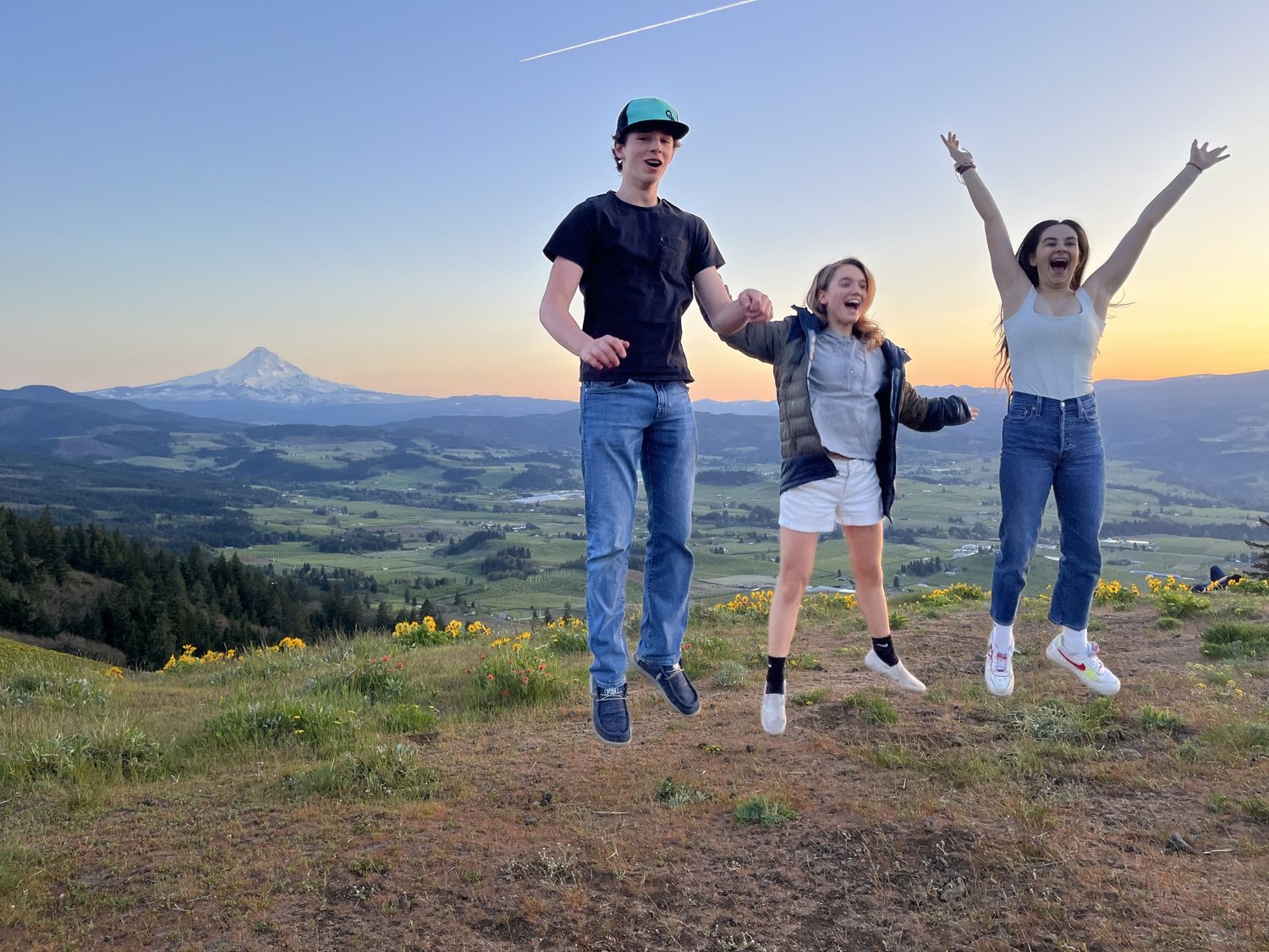 Three students jumping for joy on a hilltop at sunset, with Mount Hood behind them