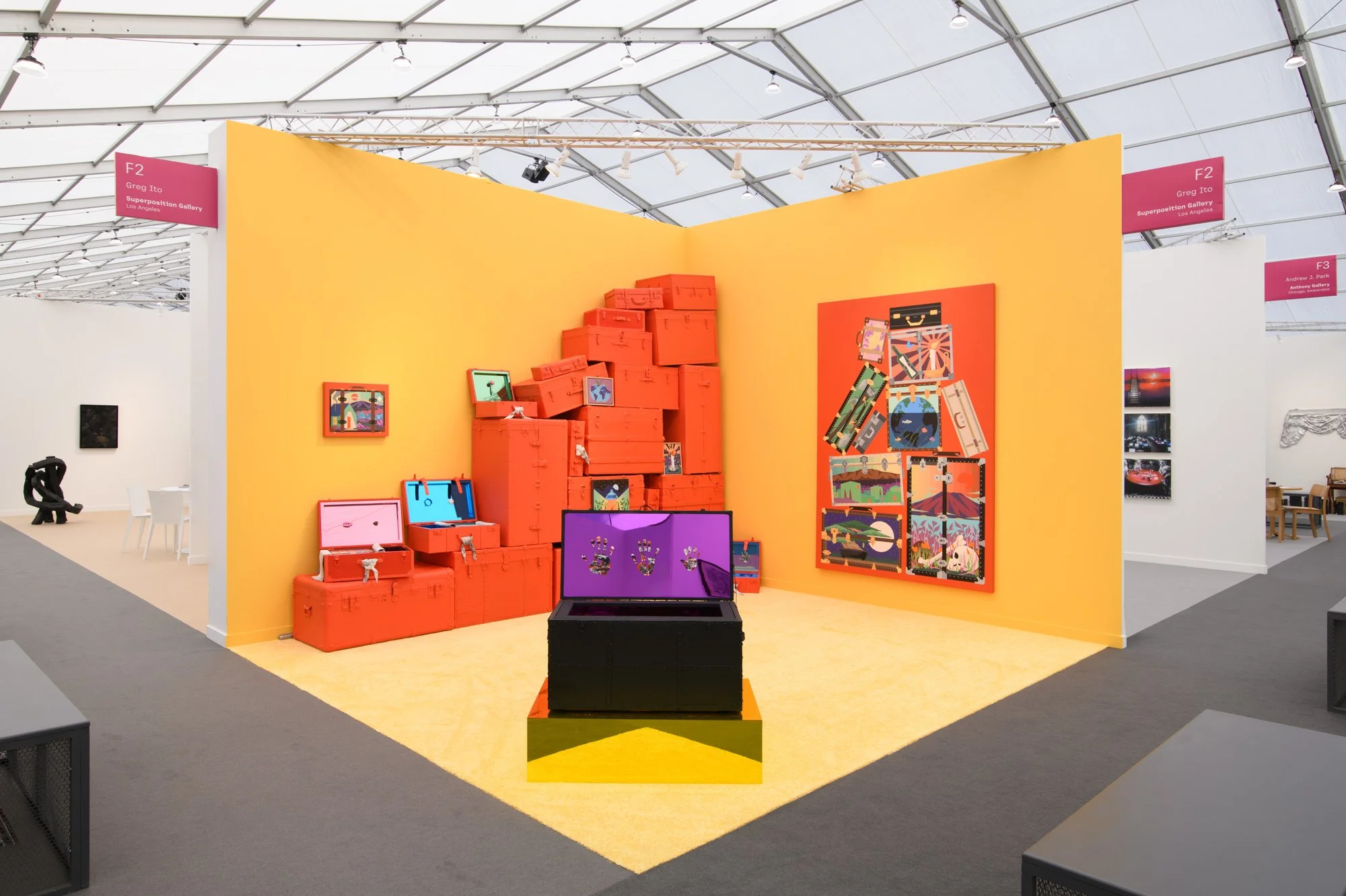 An art gallery exhibit with vibrant yellow walls displaying colorful paintings, red suitcases arranged in a pyramid, and a laptop showing jewelry designs, within a large industrial-style space with a high ceiling and bright lighting.