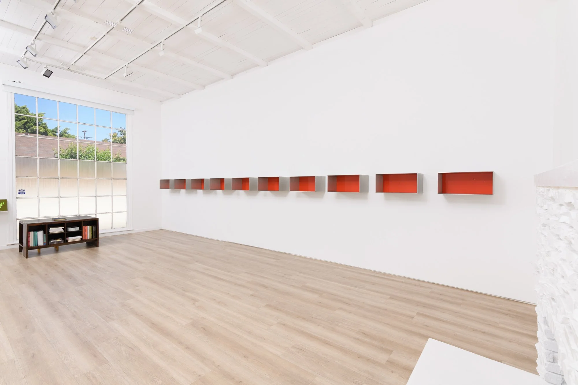 An empty art gallery with white walls, a large window, light wood flooring, and a row of small orange and gray wall-mounted artworks.