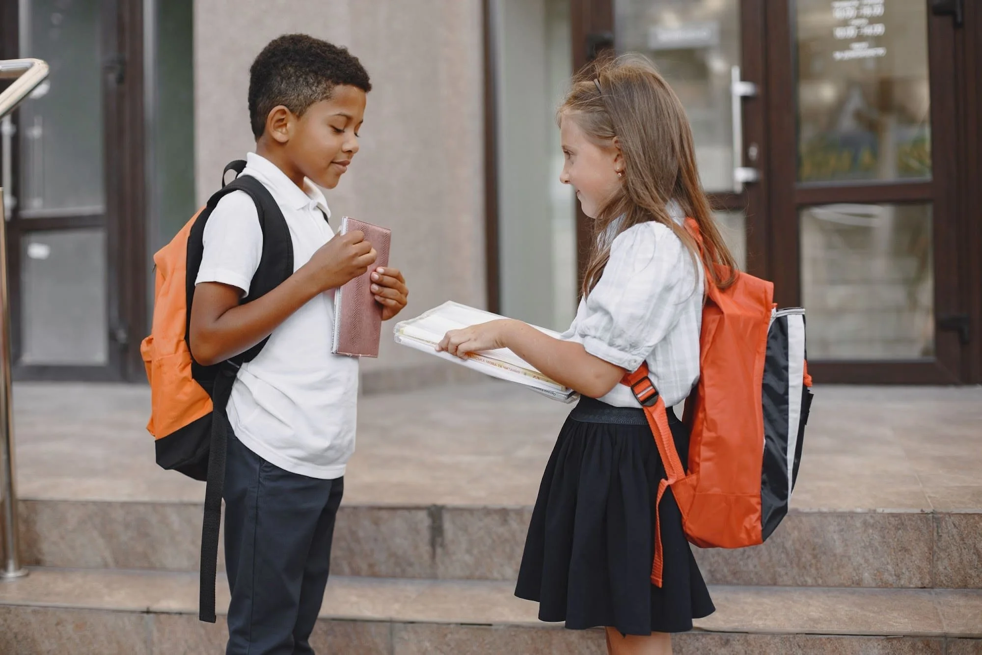 Two primary school children with backpacks standing close together outside school, sharing a moment, illustrating empathy between classmates.