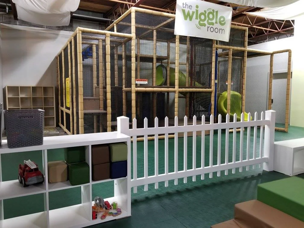 The Wiggle Room is unique in its design and intention. We want children and adults to feel comfortable and at their best. Thus, one of the greatest compliments we hear is: Your space is very well thought out. It’s in the feel and the details . And it’s true! We have consciously combined many different elements to bring The Wiggle Room to life. Starting from the top, we simulated sunlight by trading the harsh fluorescent lighting for 5,000 Kelvin LED bulbs. Plus, we made sure to bring in as much natural light as possible by selecting a space with floor to ceiling, South facing windows. Next up, we draped white sheets (that move with the HVAC) from the ceiling to suggest clouds and absorb sound. In keeping with our natural, sensory-friendly priorities, we chose muted wall colors, earth tone and recycled plastic toys, and a grass-green padded floor mat. We are also thoughtful about sonic stimulation; our classical music playlist enriches little brains without overtaking the play. All these choices, combined with all the time we spend here with our own children, make this place a true extension of our home. We welcome you to come experience The Wiggle Room!