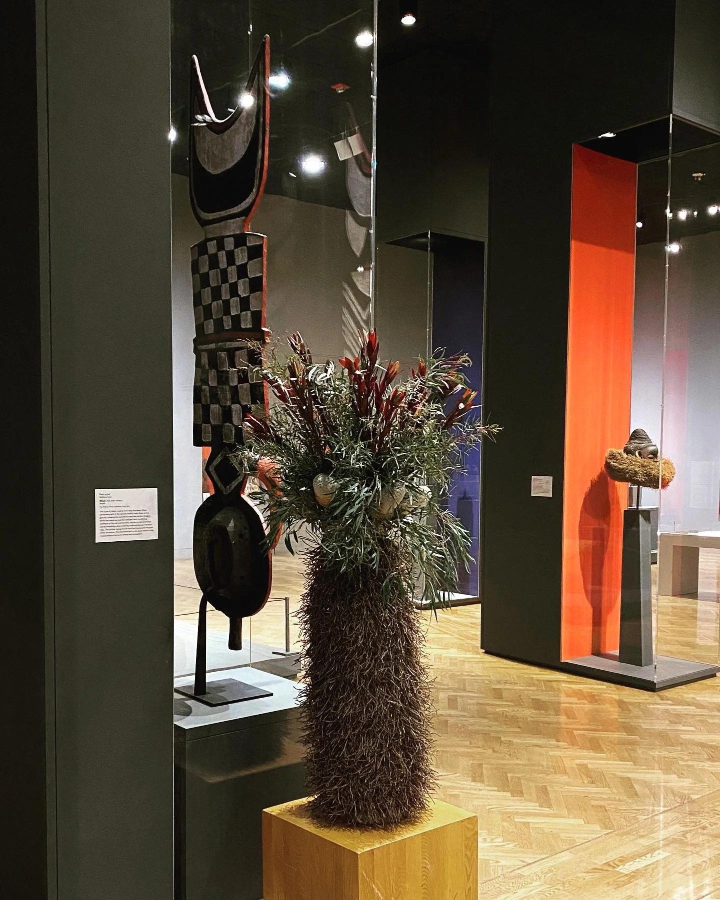 A tall wooden mask sculpture in a museum enclosed in a glass case with a wooden pedestal beside it featuring a floral arrangement.