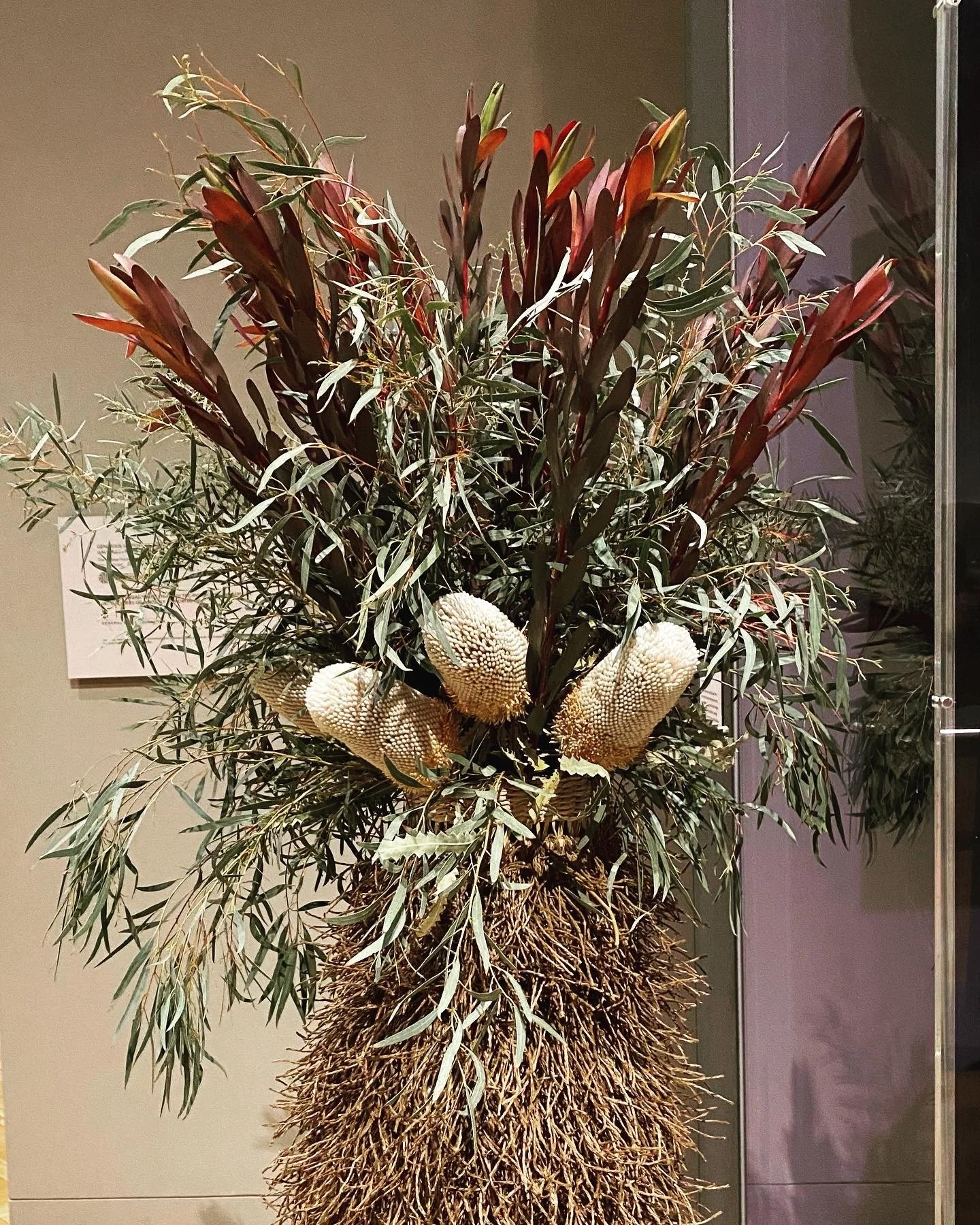 A tall arrangement of florals including agonis leaves and large white banksia flowers at the base housed in a knotted fringed tall vase.