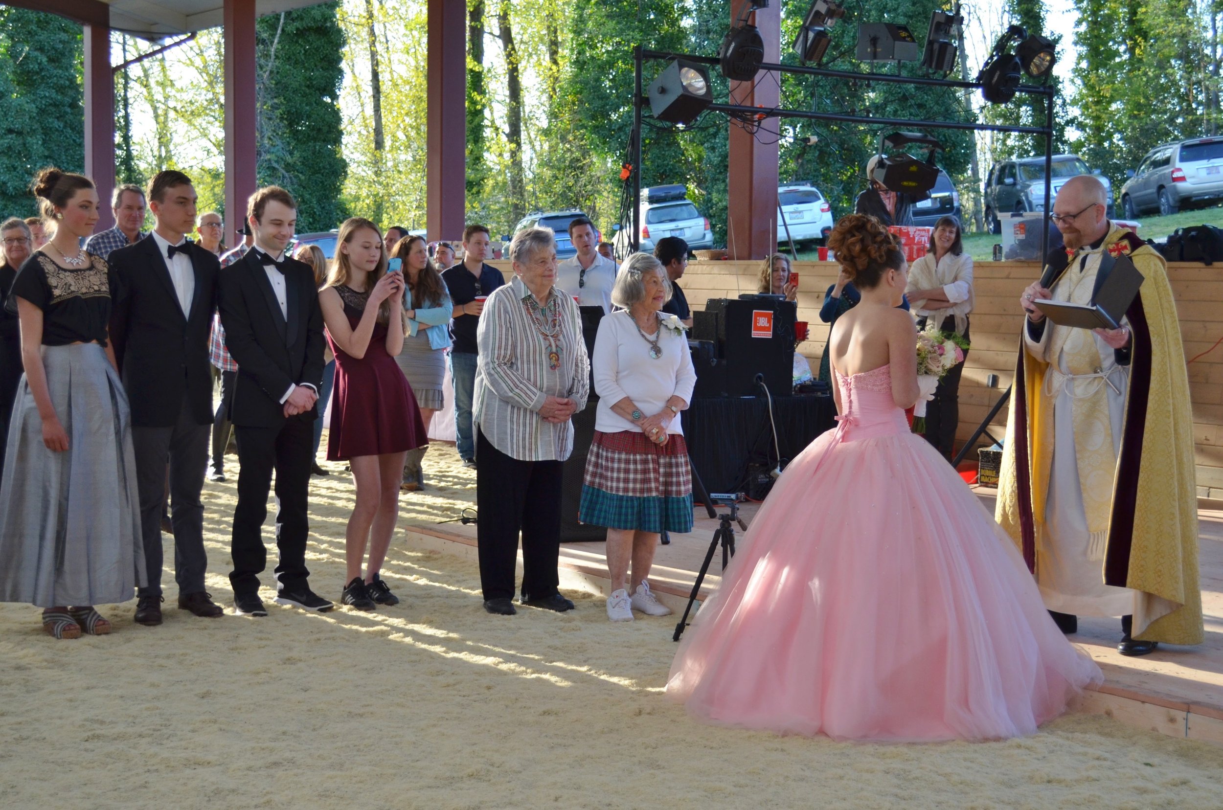 A ceremony with a young woman in a pink ball gown receiving an award or recognition from a man dressed in regal attire, with an audience of young adults and elderly women watching.